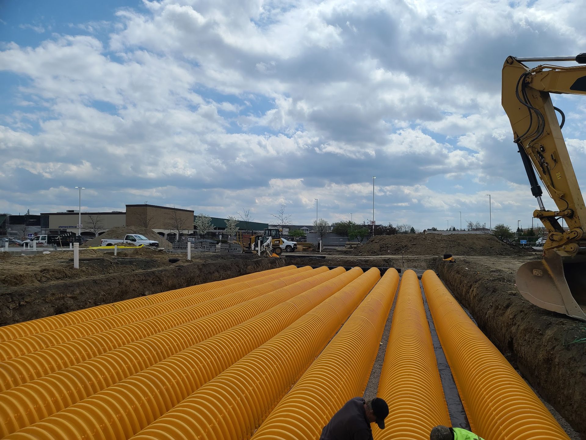 Yellow corrugated pipes in a trench at a construction site; excavator and buildings visible under a cloudy sky.