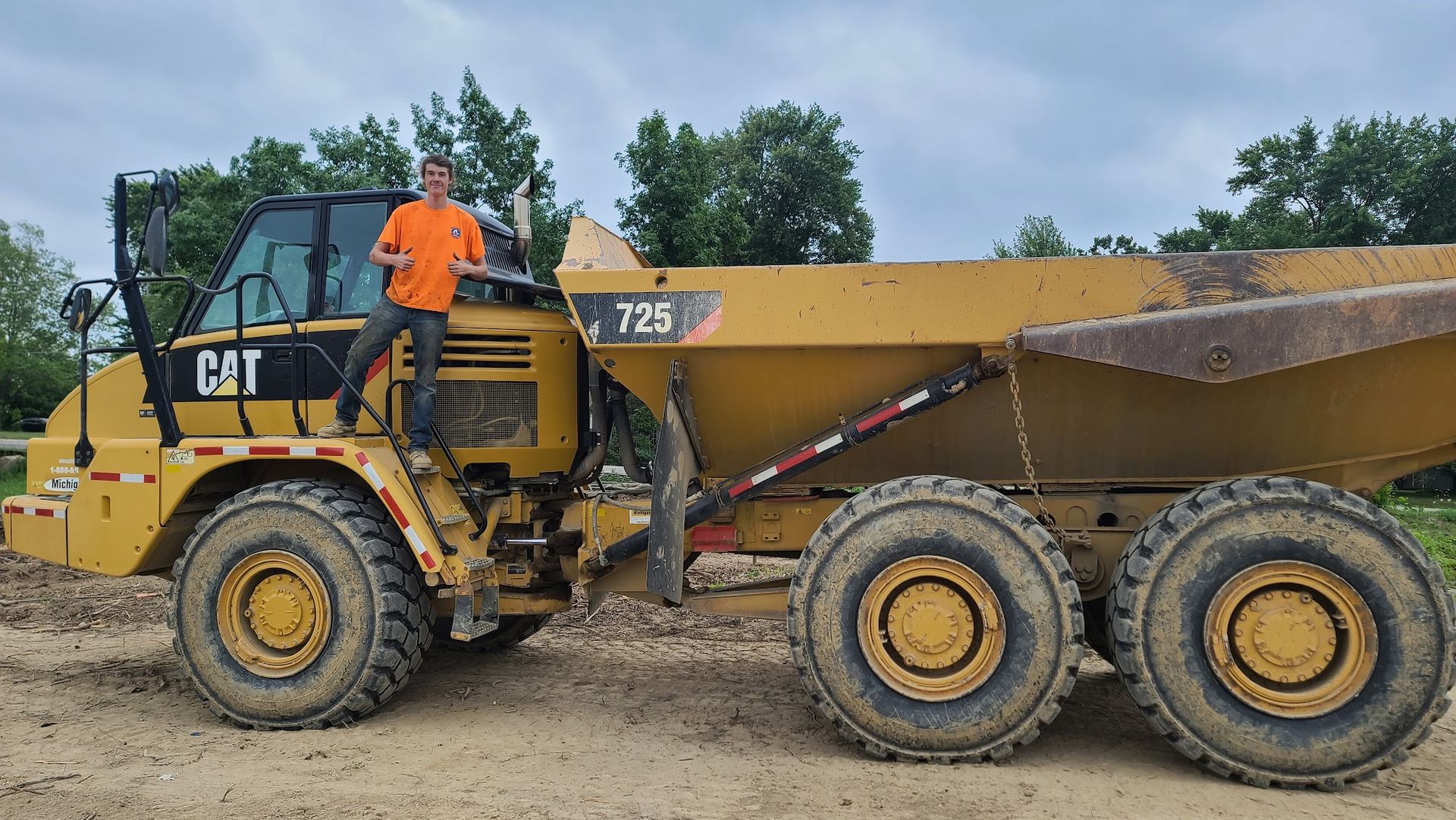 Man in orange shirt standing on yellow Caterpillar 725 dump truck. Outdoors, trees in background.