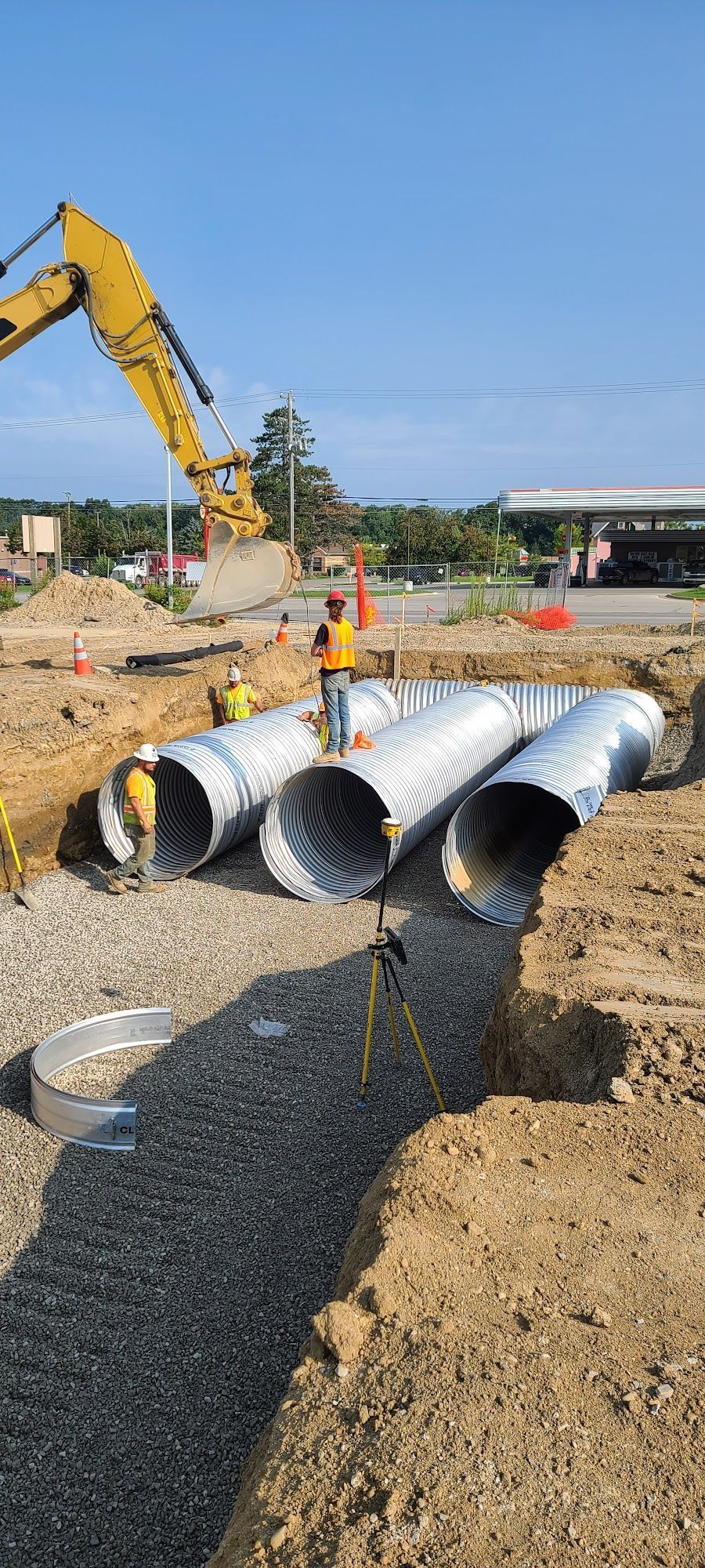 Construction site with large corrugated metal pipes being installed. Workers and excavator visible.
