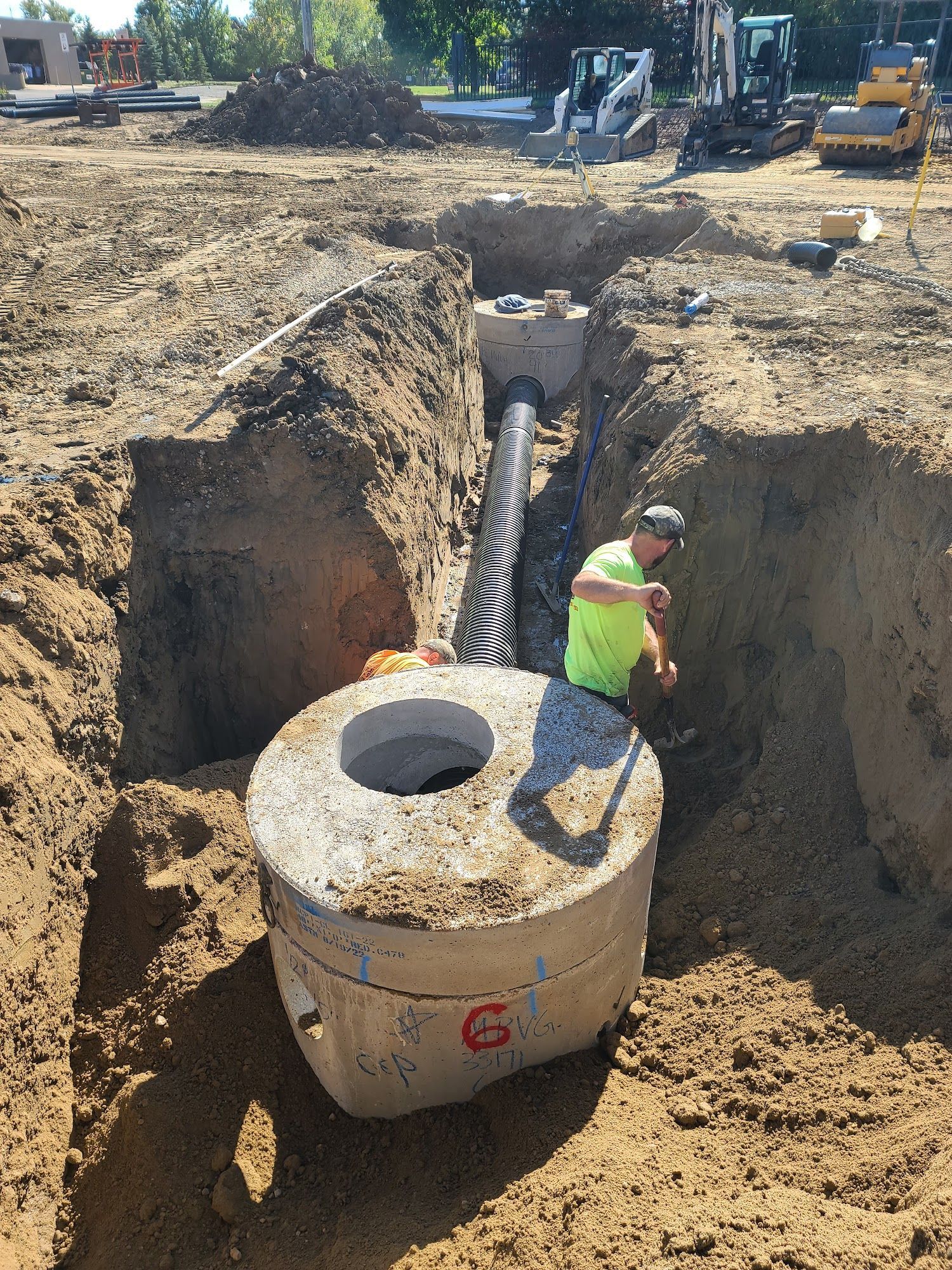 Construction site with a worker installing a concrete sewer structure, surrounded by trenches and machinery.