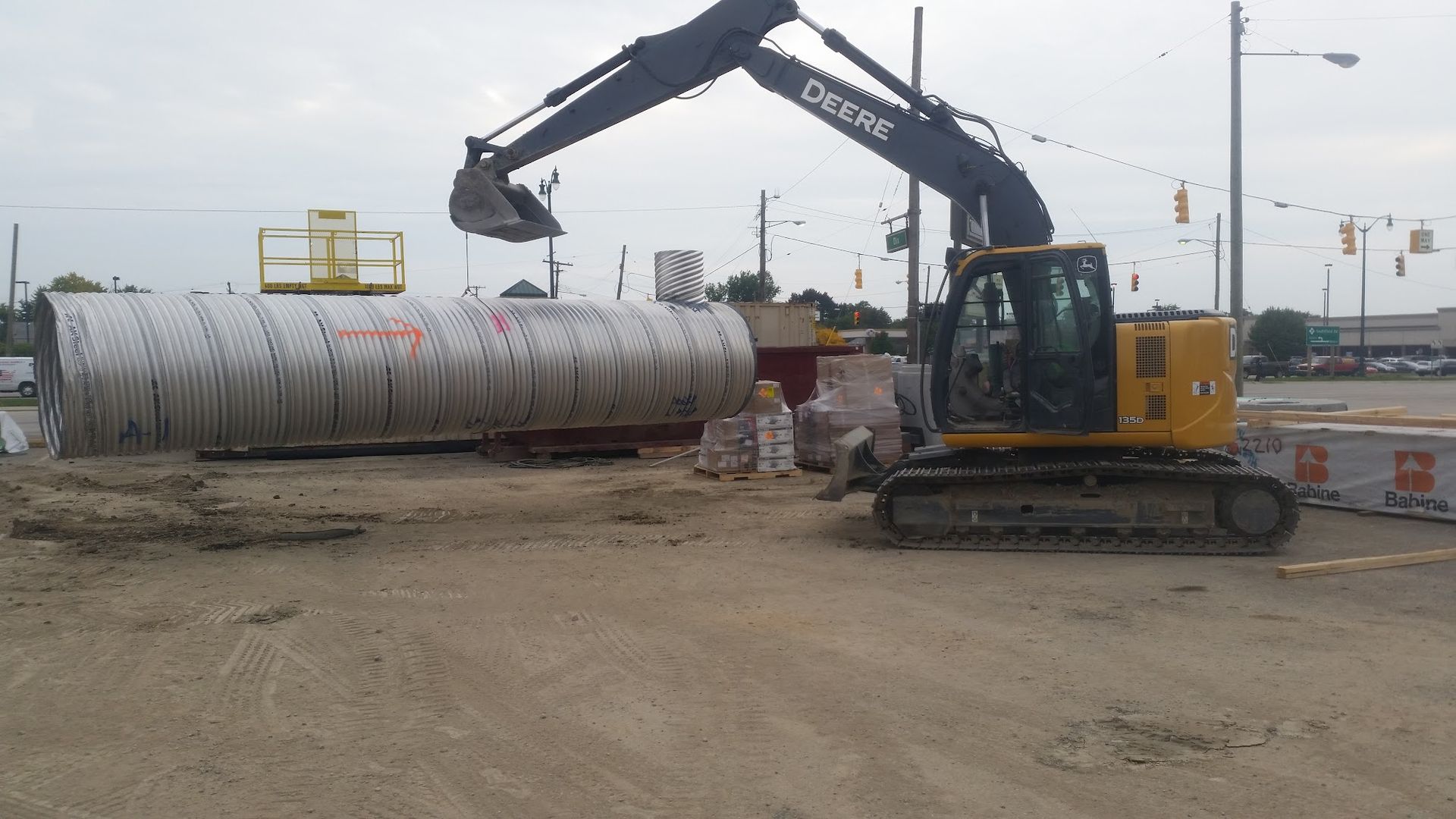 Excavator with bucket lifting a large concrete pipe at a construction site.