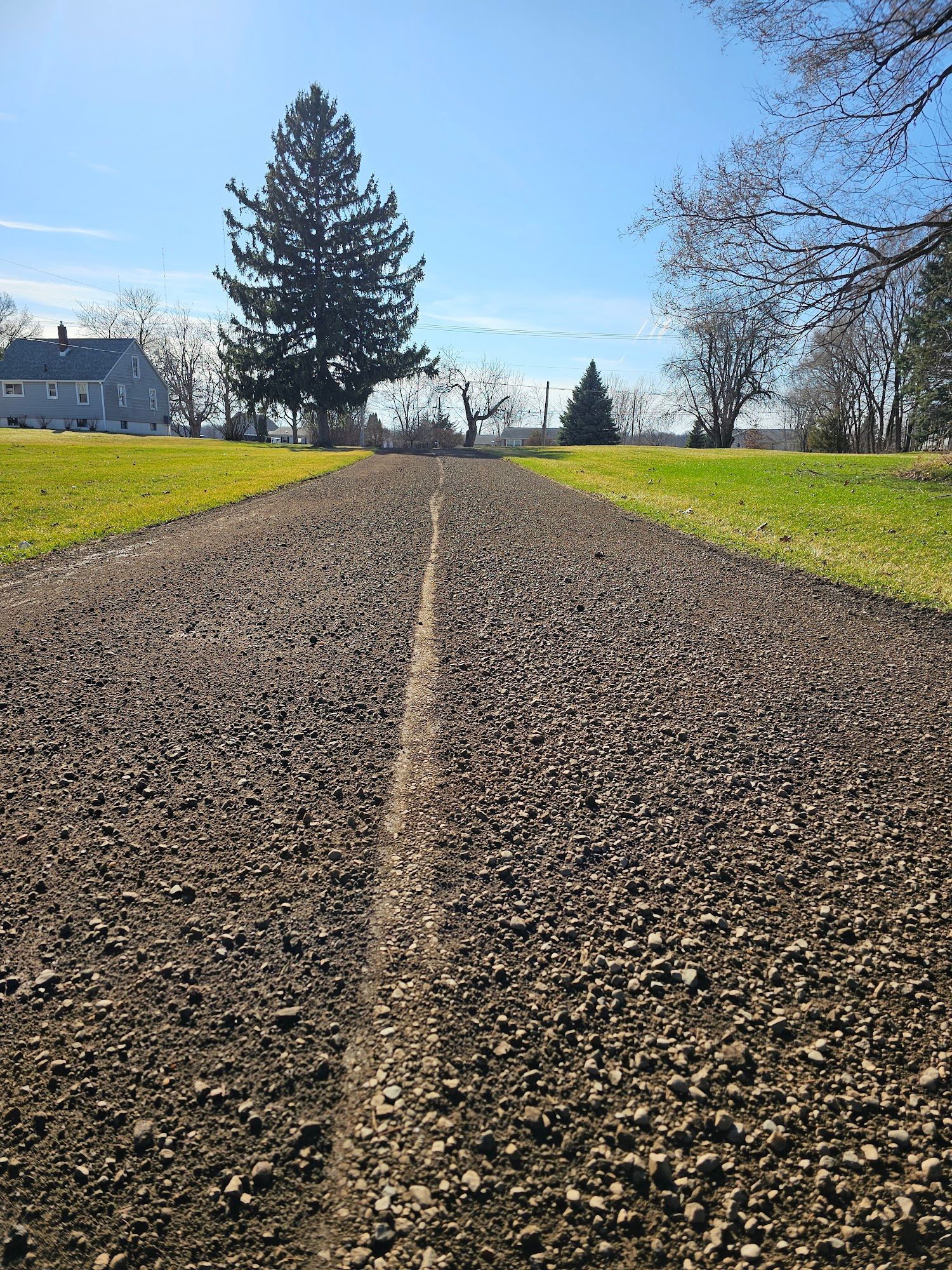 Gravel path with a white line down the center, flanked by green grass. Buildings and trees in the distance.