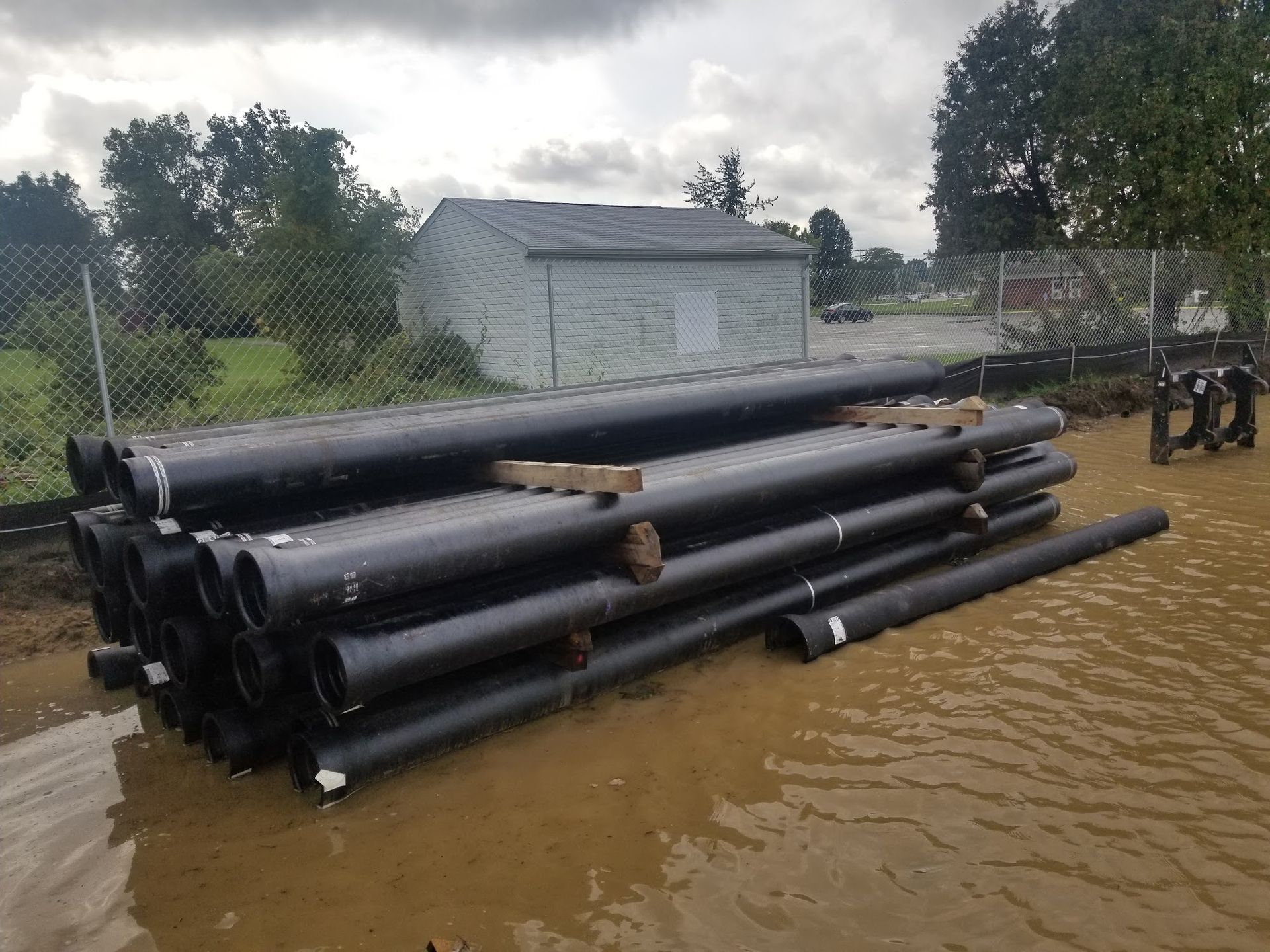 Black pipes stacked in a muddy area near a gray building and a chain-link fence.