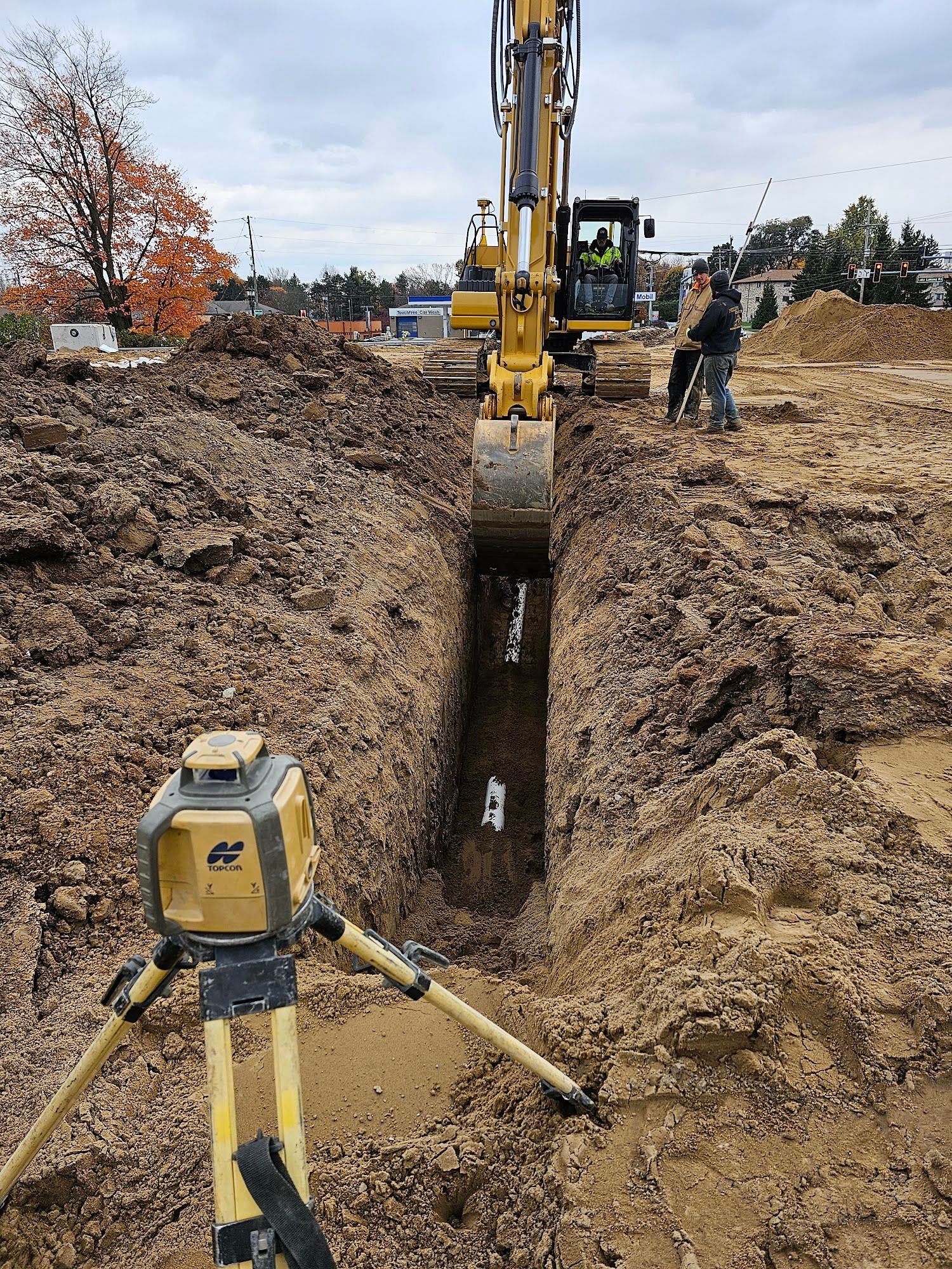 Yellow excavator digging a trench, with a laser level in the foreground and a worker nearby.