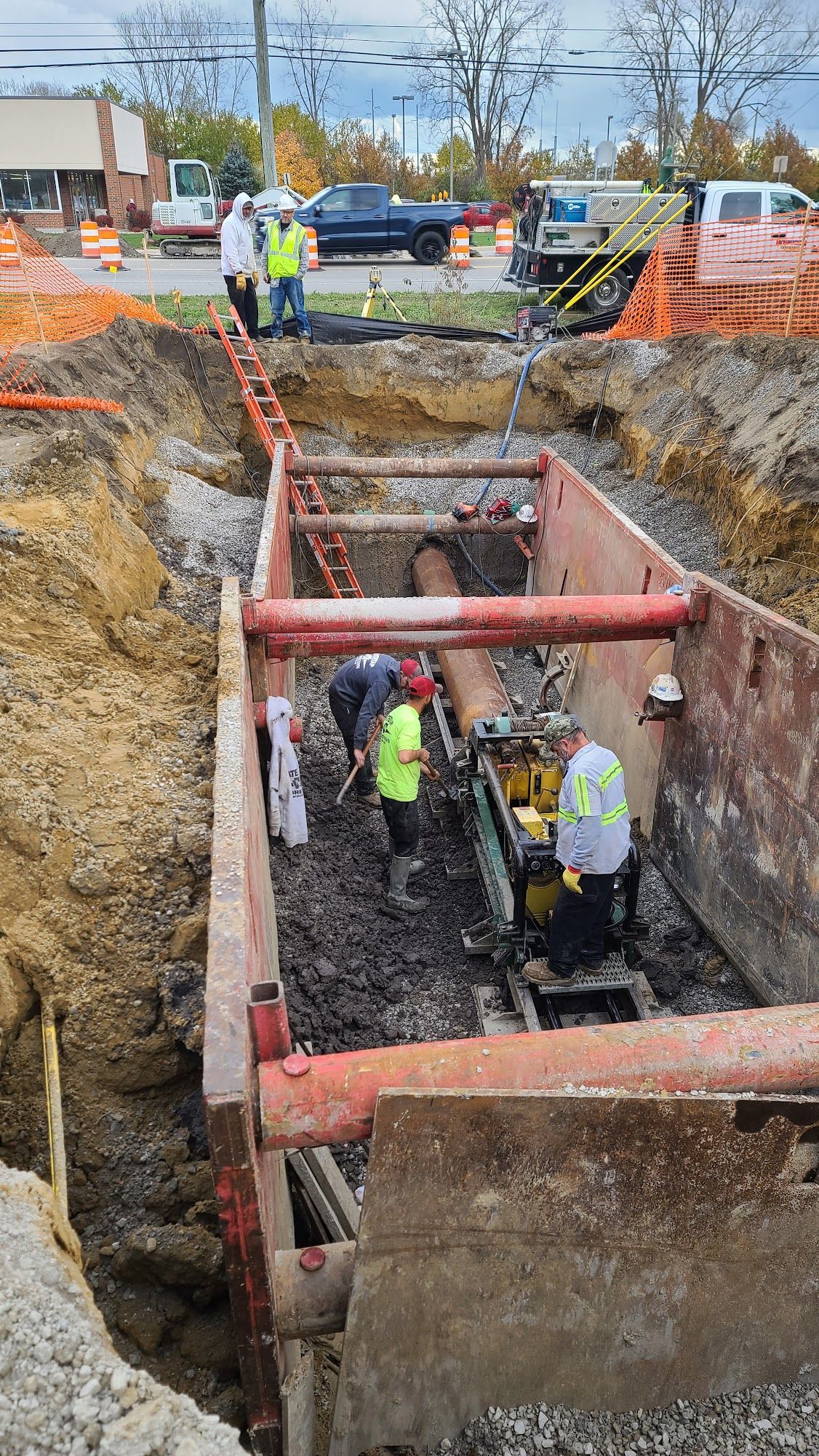Workers in a deep construction trench installing or repairing underground pipes. Orange safety barriers surround the site.