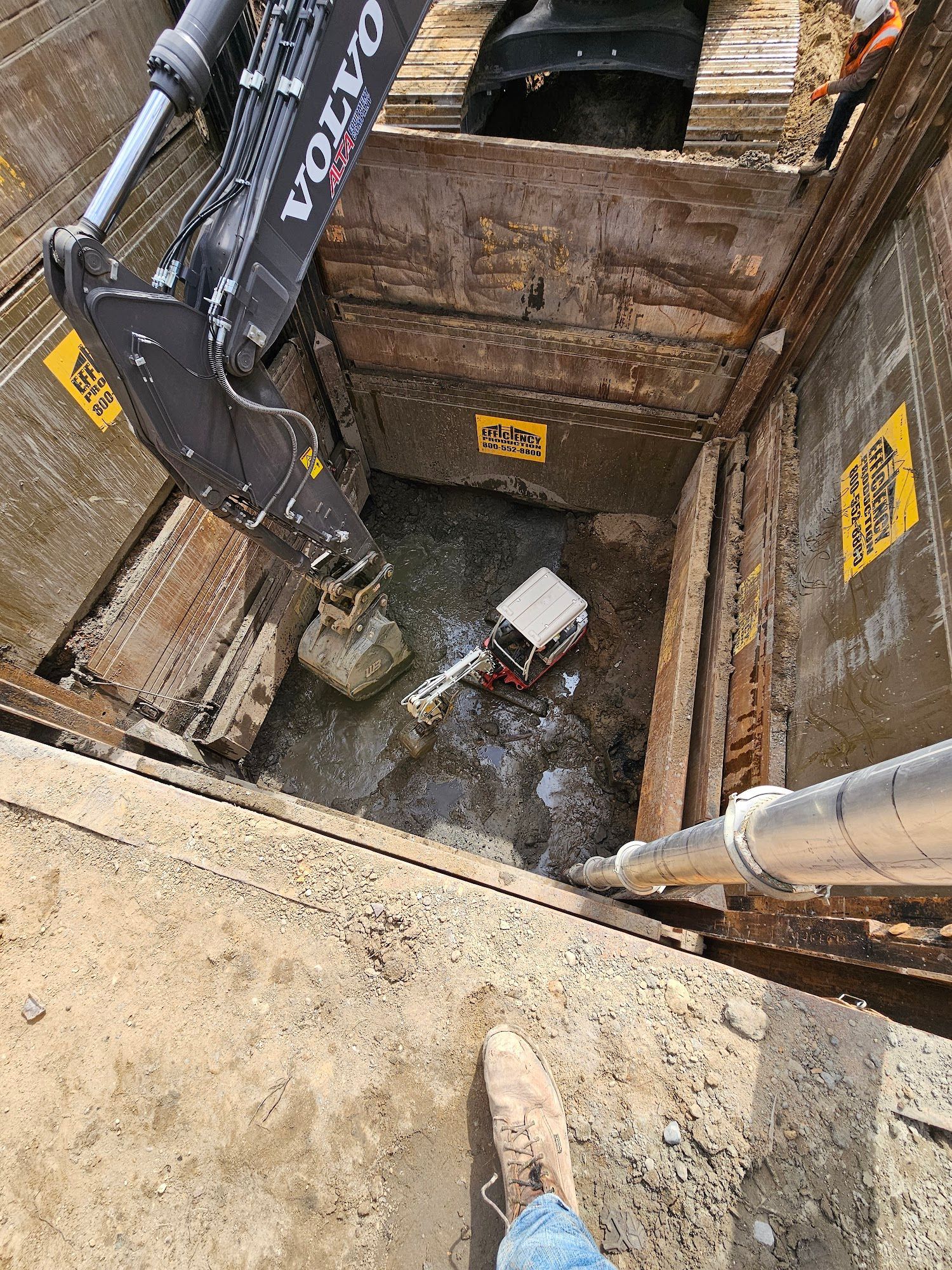 Excavator in a trench, removing debris. Metal shoring walls. Man's feet in foreground.