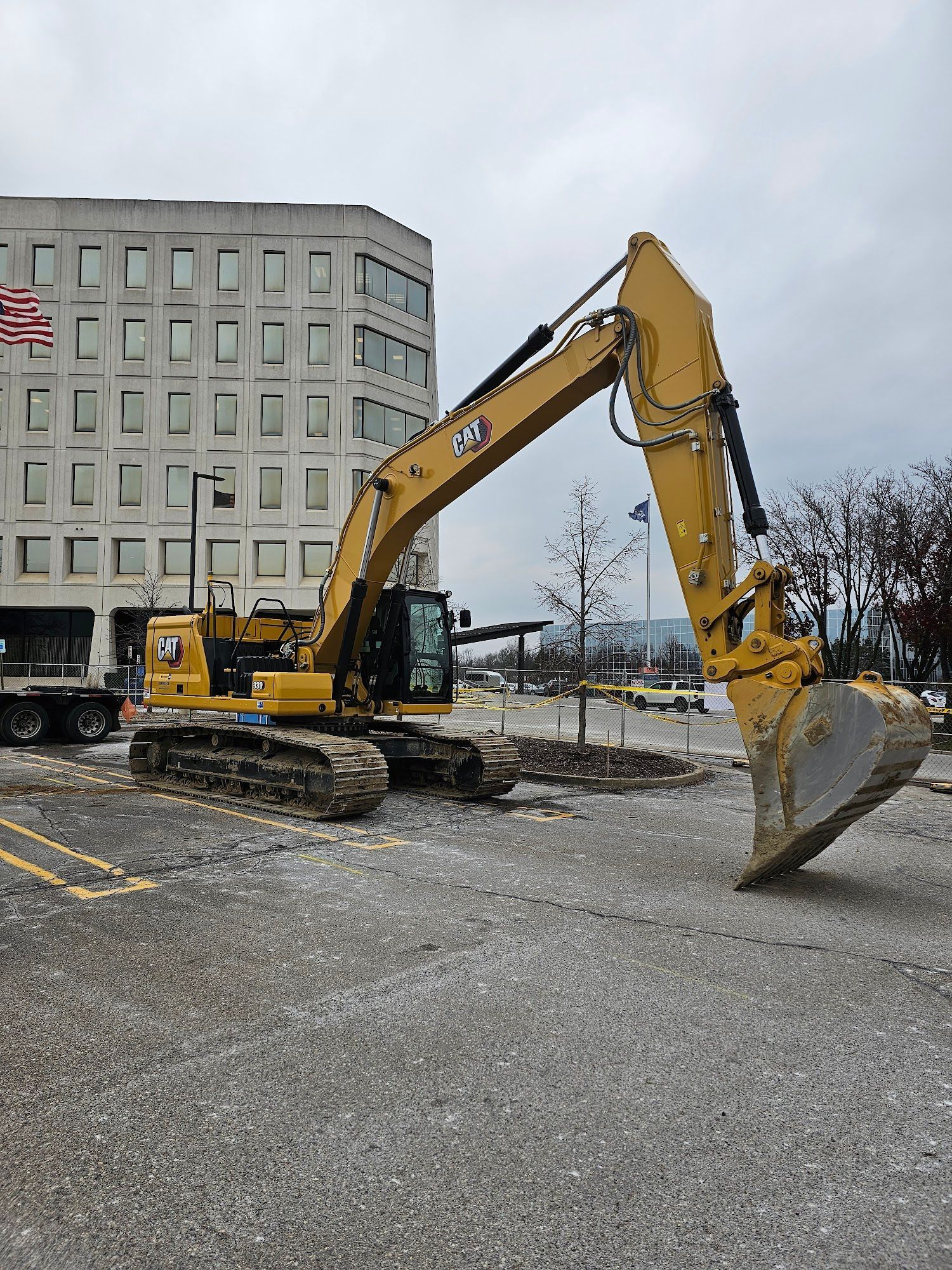 Yellow Caterpillar excavator in a parking lot, near a large building with an American flag.