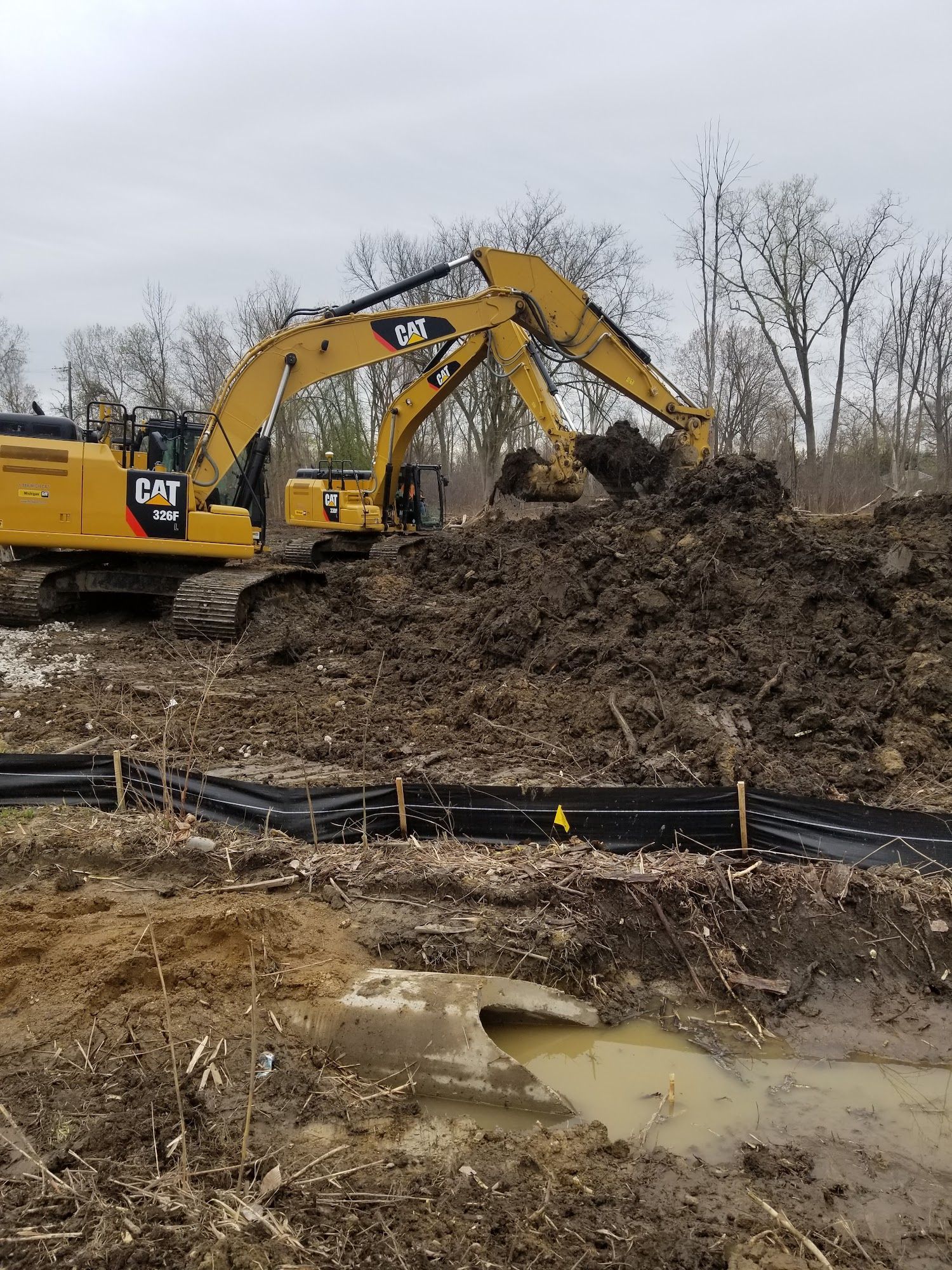 Yellow excavator digging dirt on a muddy construction site. Black pipes and a puddle visible.