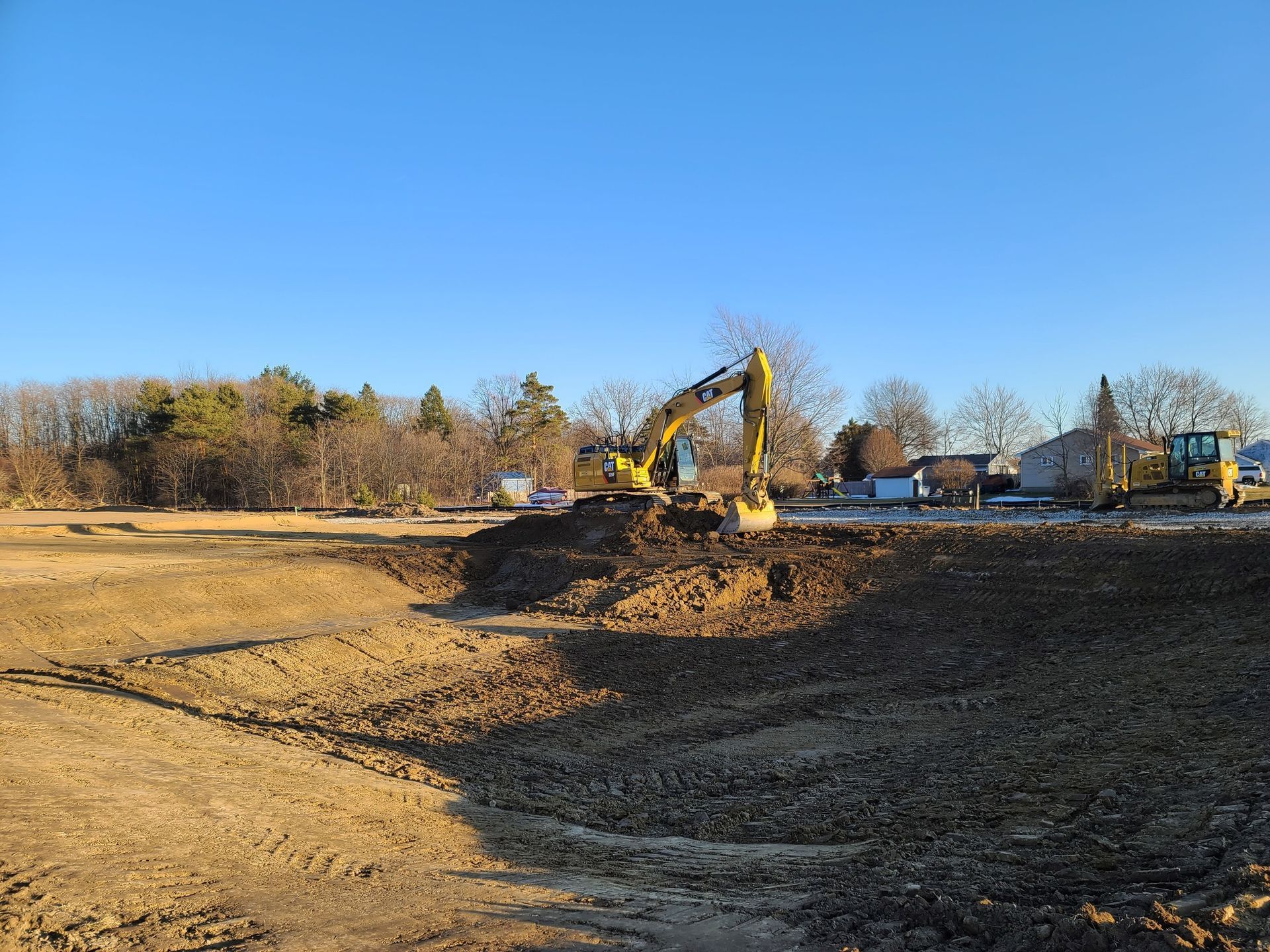 A yellow excavator digging in a large, dirt-filled construction site under a clear blue sky.