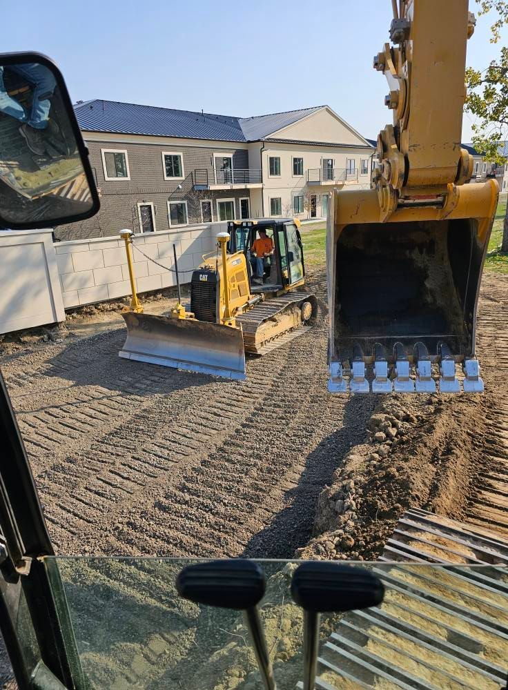 Construction site with excavator bucket, skid steer, and graded dirt beside new buildings