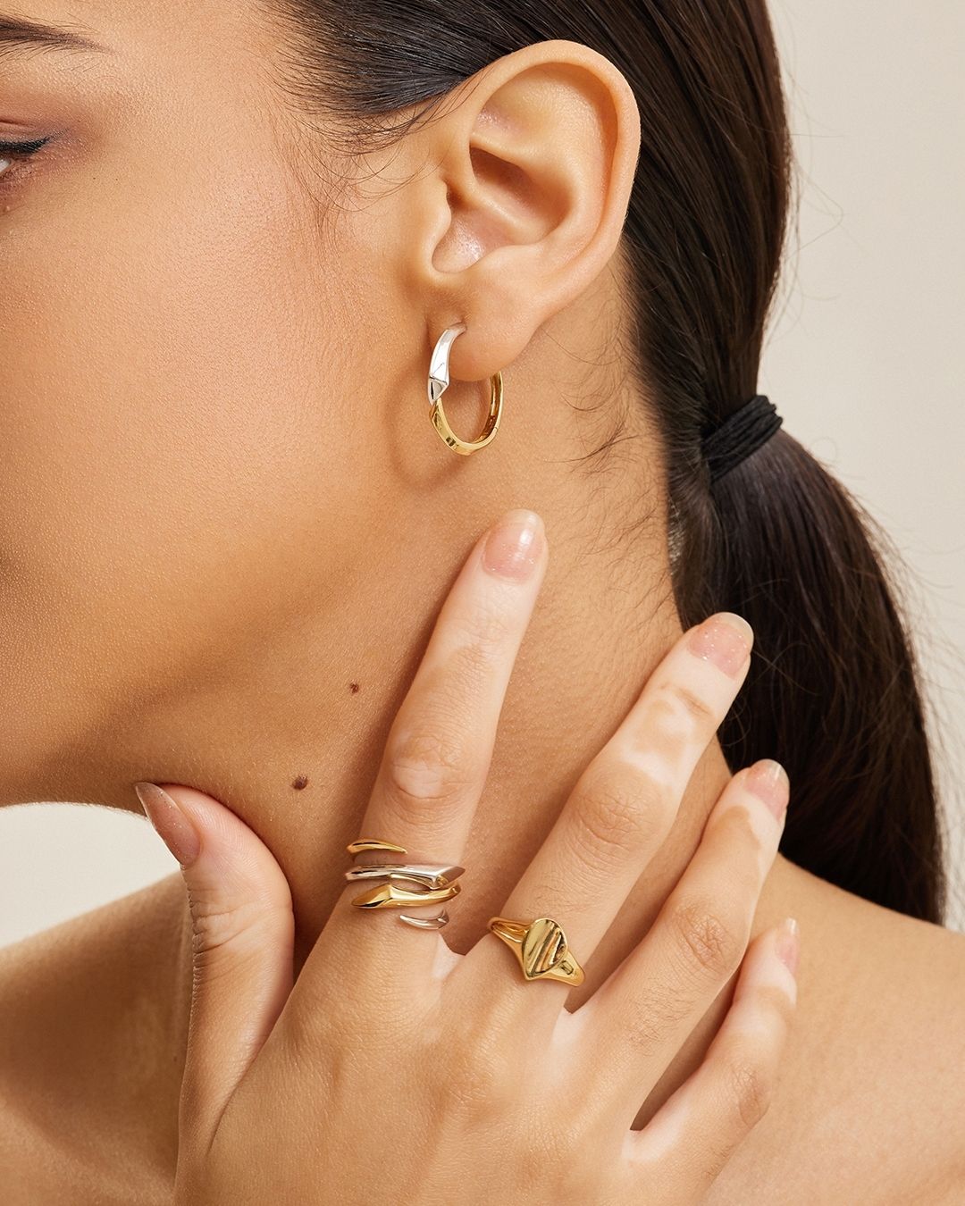 Femme portant des boucles d'oreilles créoles et des bagues en or, touchant son cou avec sa main.