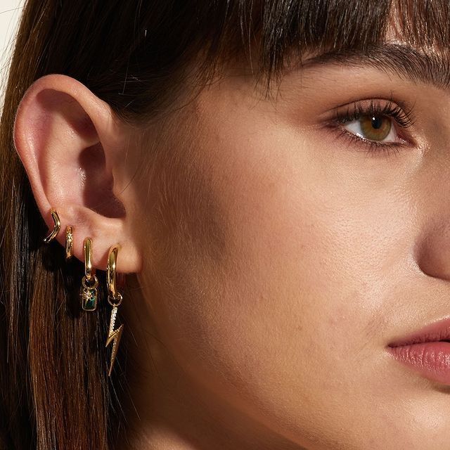Oreille de femme avec plusieurs boucles d'oreilles en or : un éclair, une pierre précieuse et des créoles. Cheveux bruns, peau claire.
