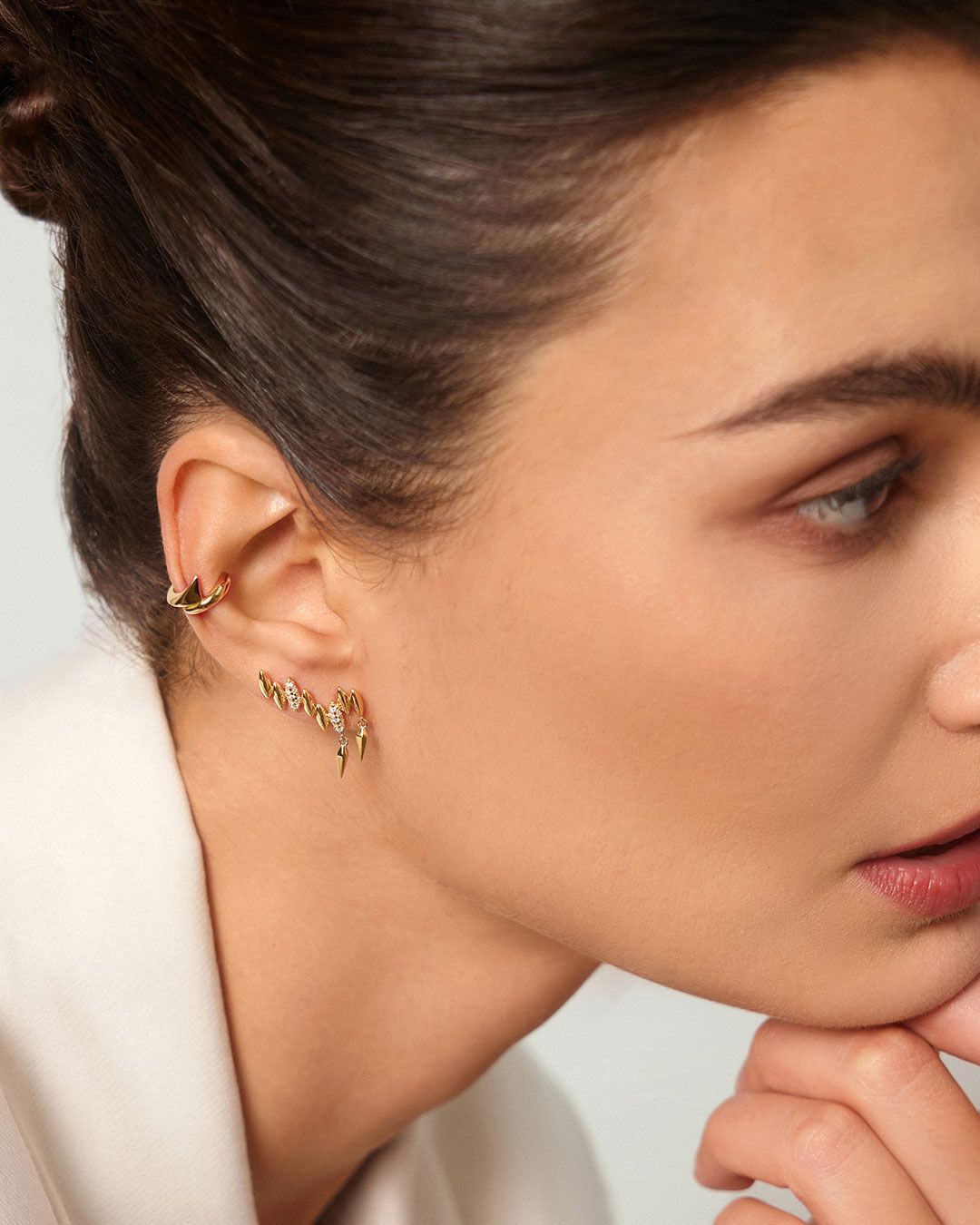 Femme portant des boucles d'oreilles en or, regardant vers la droite, vêtue d'une veste blanche. Ses cheveux sont relevés.