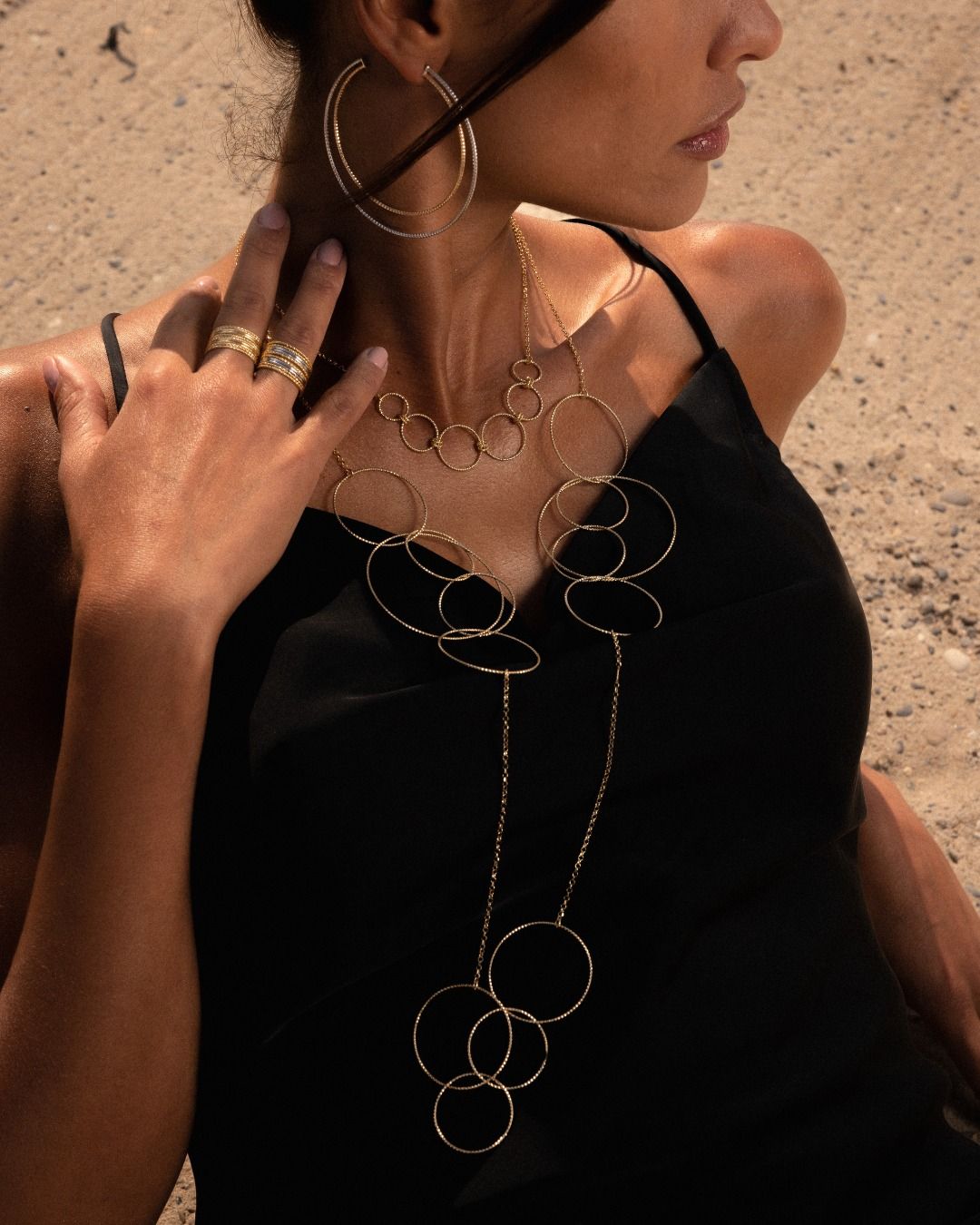 Femme portant des bijoux en or : boucles d'oreilles créoles, bagues et collier, robe noire, soleil.