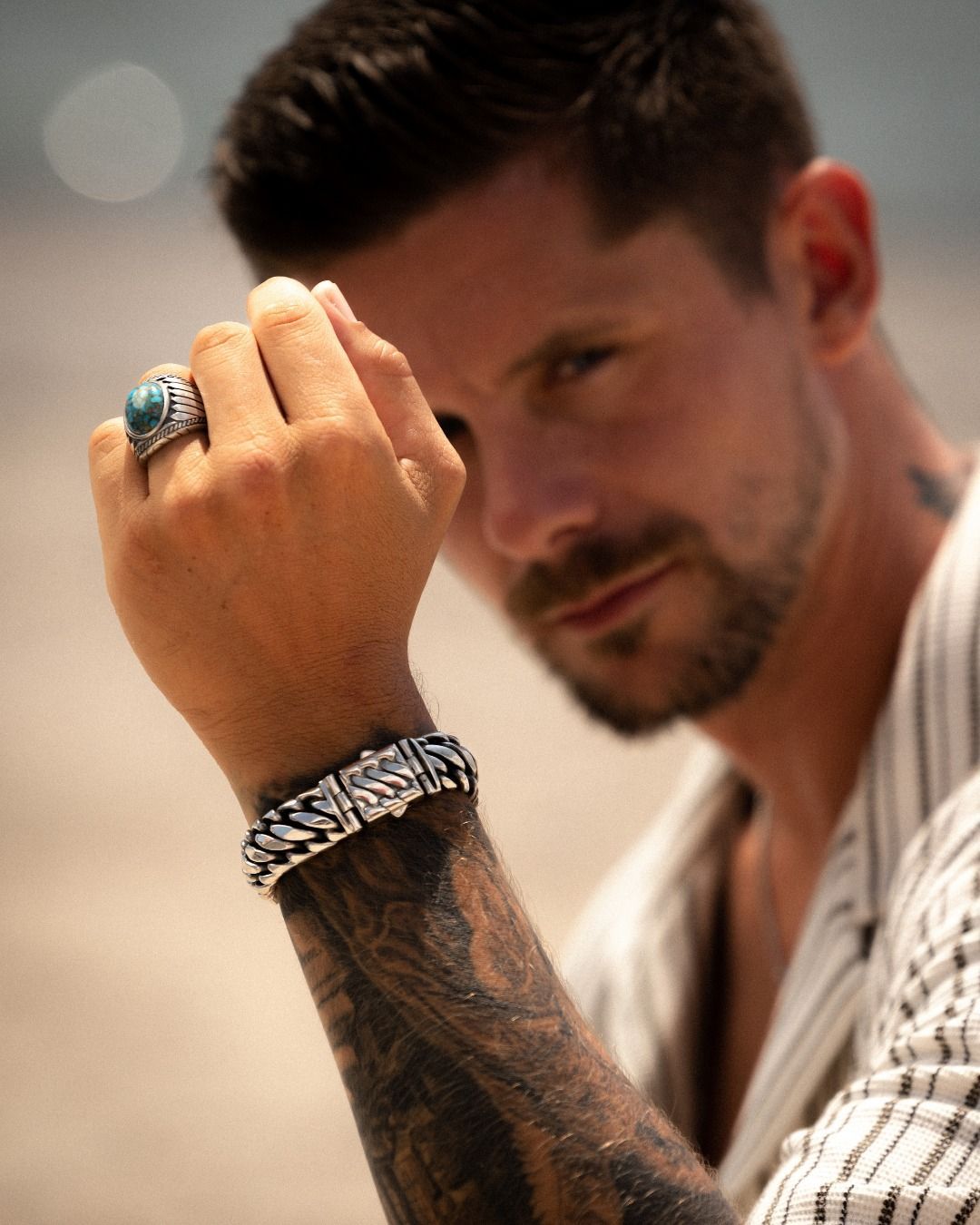 Homme avec bague et bracelet, regardant la caméra. Tatouage au bras, fond sable, portant une chemise rayée.
