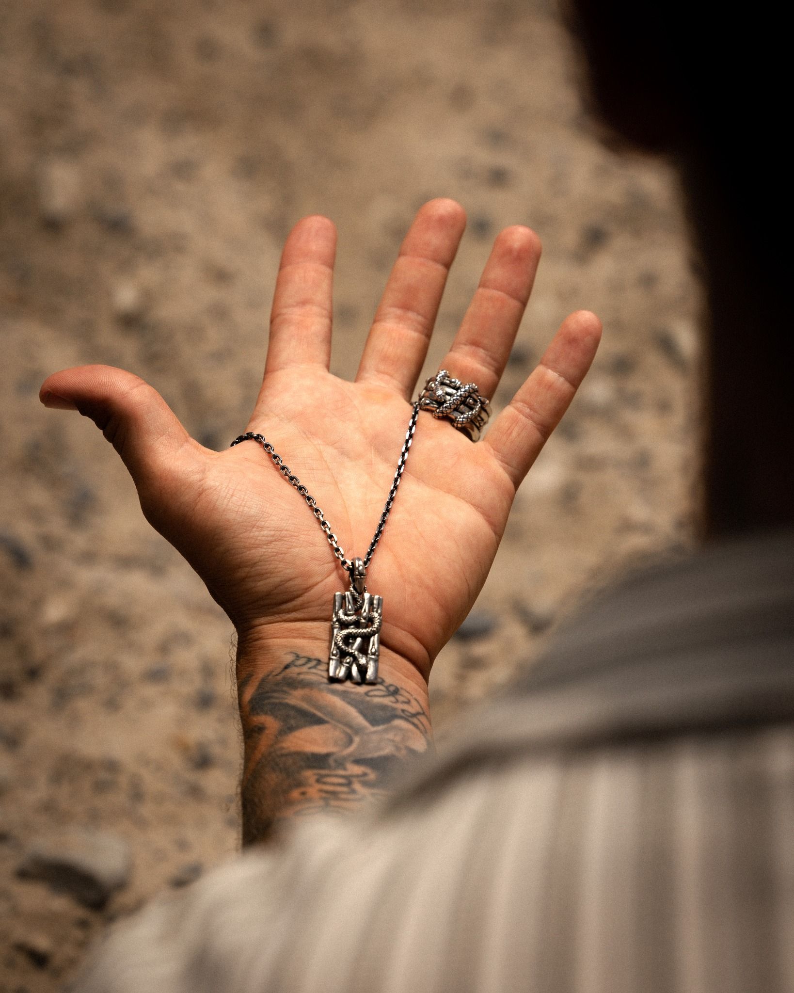 La main de la personne présente un collier et une bague en argent, un tatouage sur le bras est visible. L'arrière-plan est flou, probablement en extérieur.