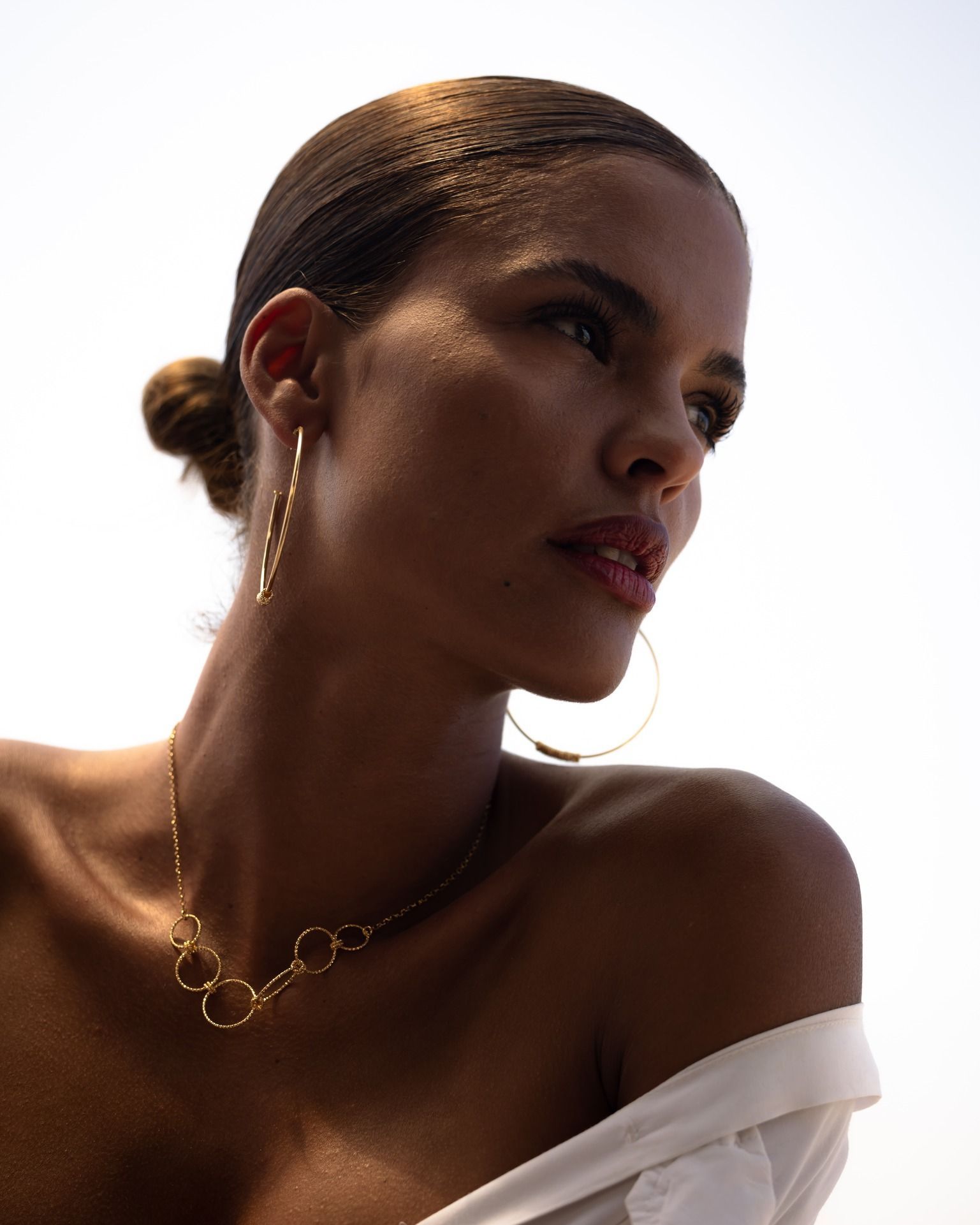 Femme aux cheveux noirs et aux bijoux en or, regardant ailleurs, chemise blanche, fond de ciel.