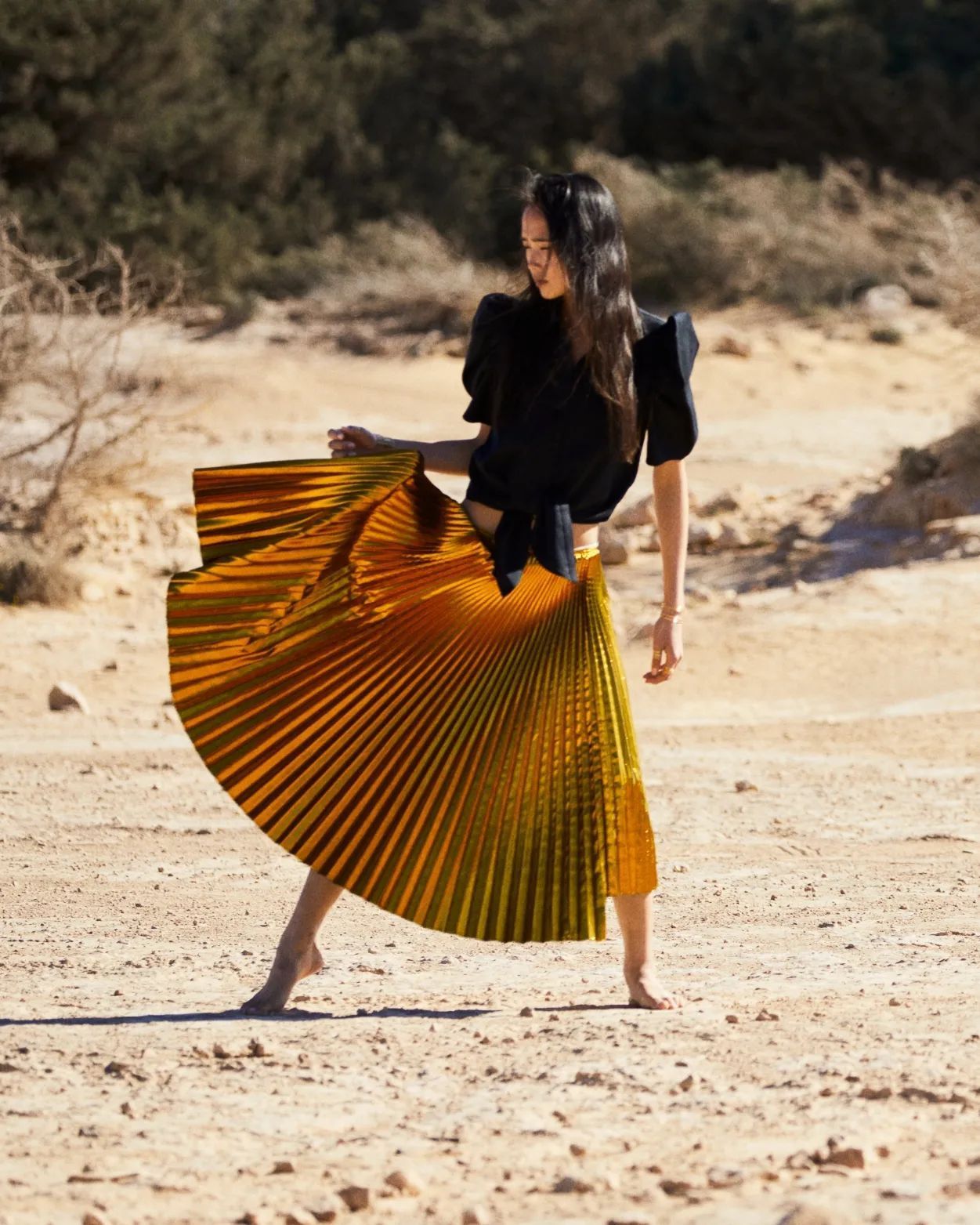 Une femme en haut noir et jupe plissée dorée danse sur un sol sablonneux.