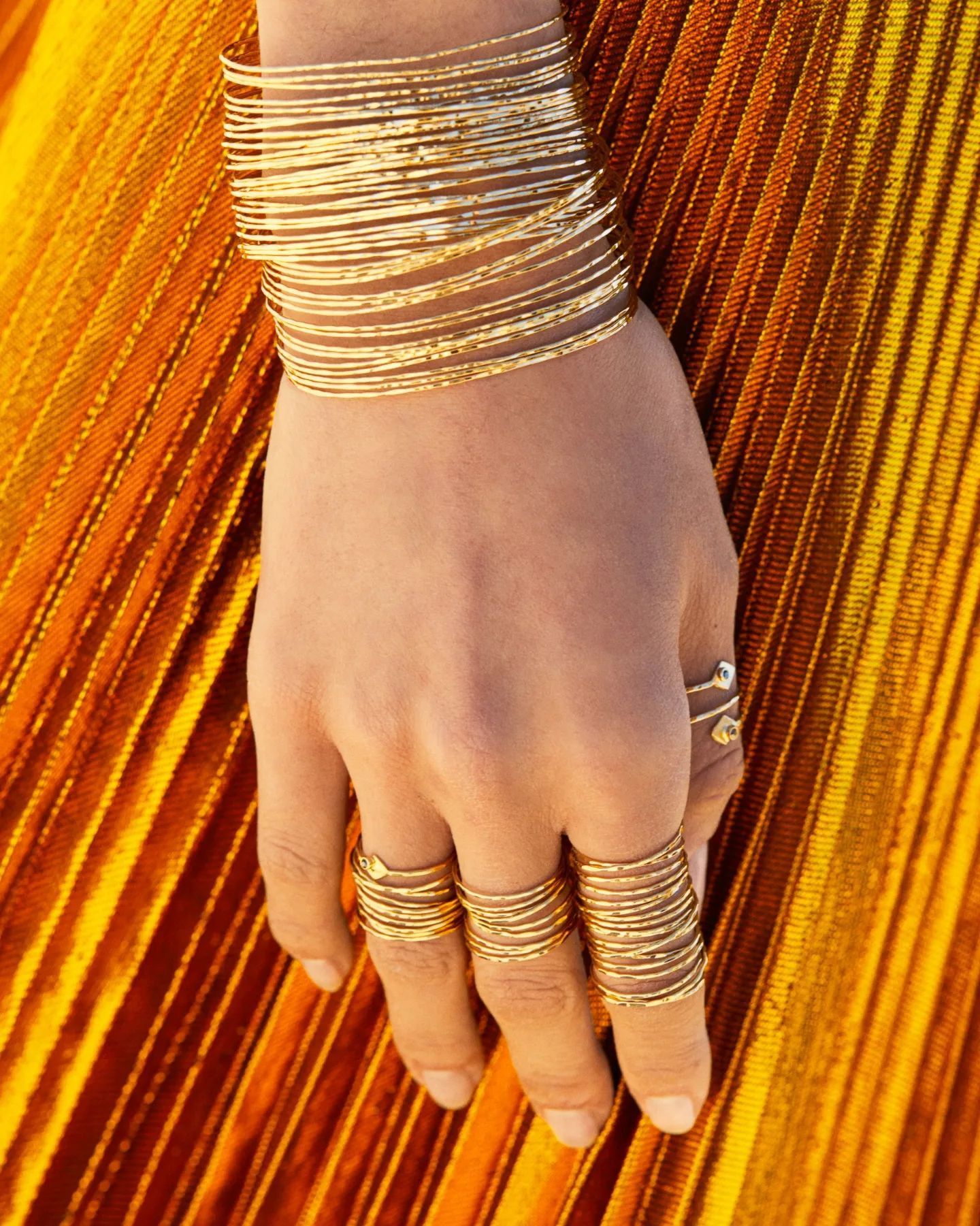 Main ornée d'anneaux et de bracelets en or, reposant sur un tissu orange plissé.