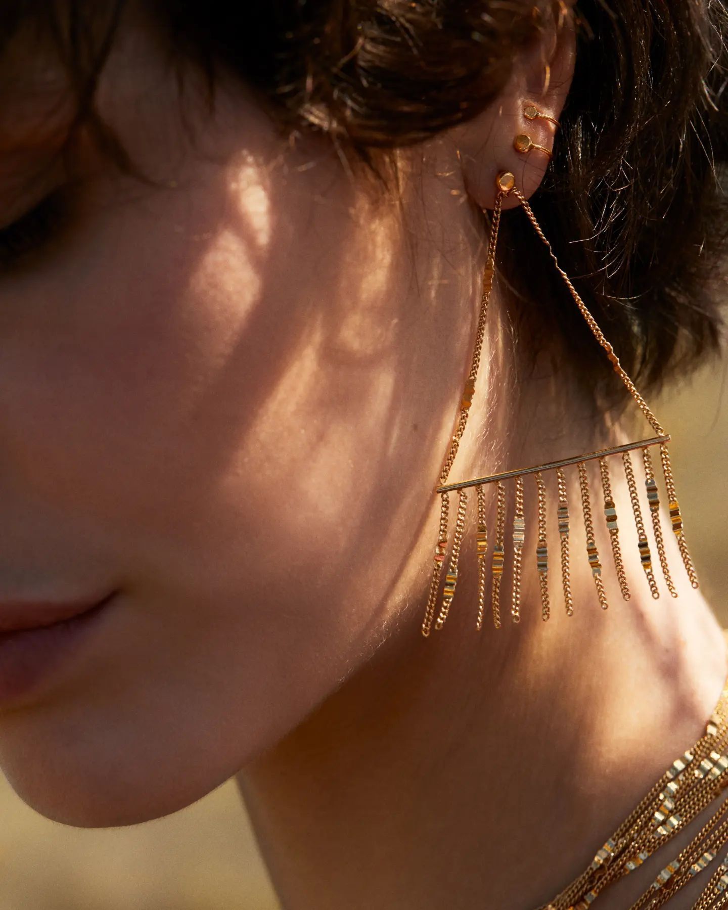 Femme portant des boucles d'oreilles à franges en or, la tête inclinée, avec la lumière du soleil projetant des ombres.