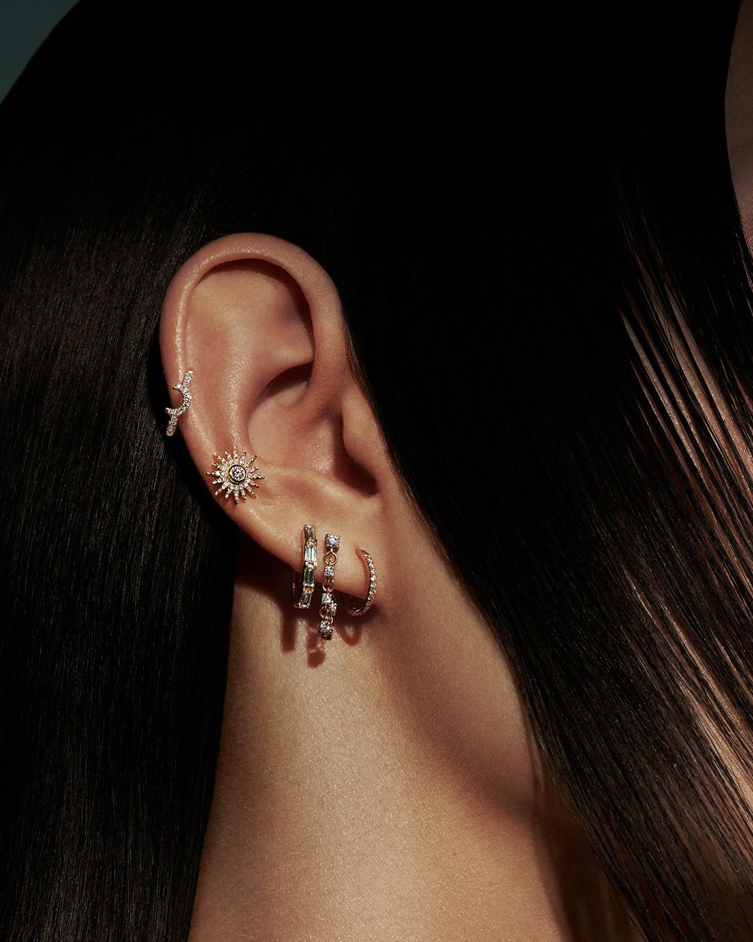 Oreille ornée de multiples boucles d'oreilles : en forme de soleil, créoles et clous. Des cheveux foncés encadrent l'oreille.