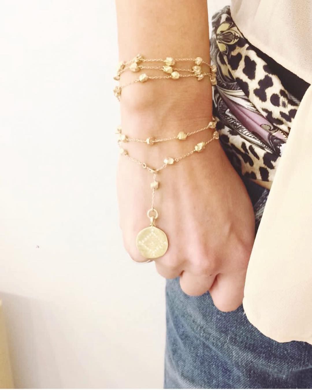 Bracelets en or superposés au poignet, avec un pendentif en forme de pièce. Une main est visible, vêtue d'un jean.