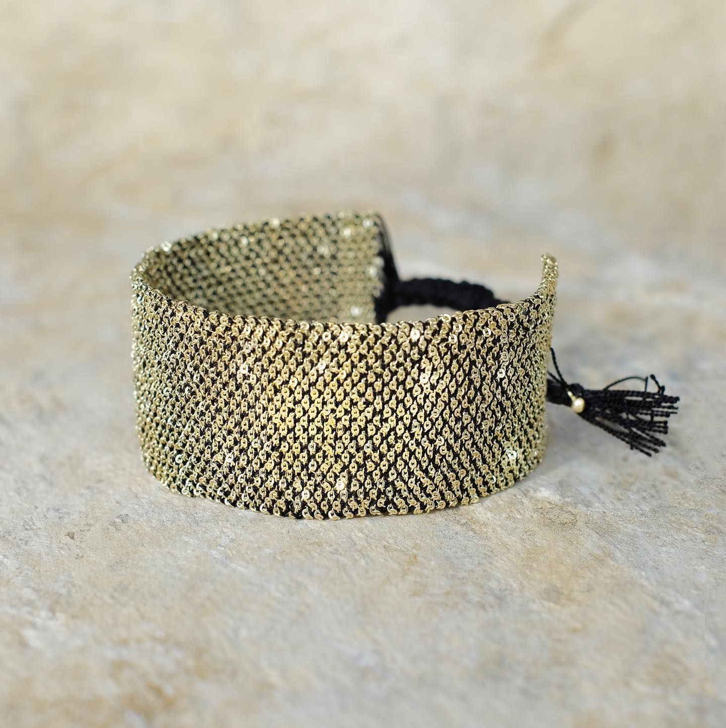 Bracelet tissé en or et noir avec un pompon noir sur une surface beige.