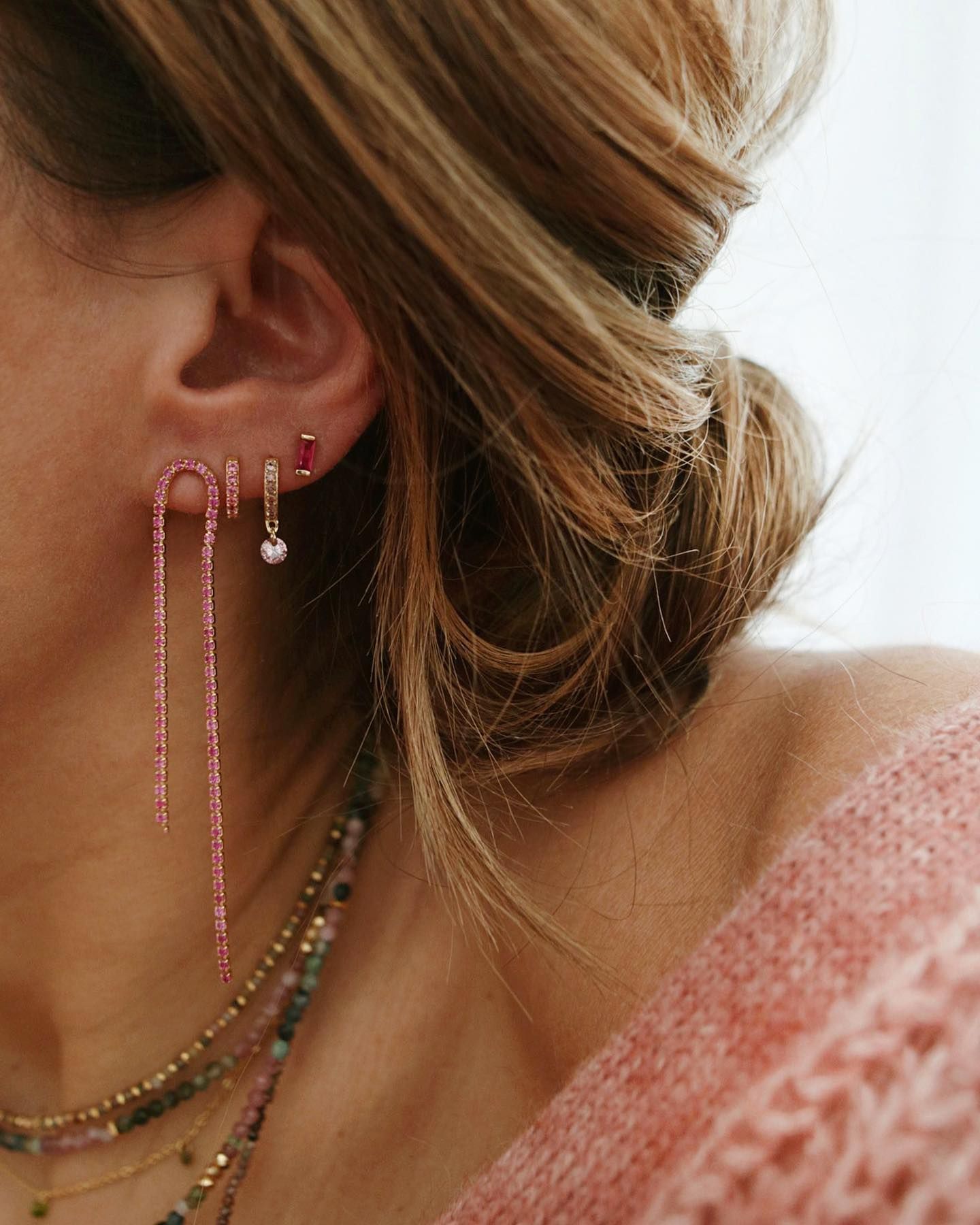 Femme avec plusieurs piercings aux oreilles portant des boucles d'oreilles roses, certaines pendantes, avec des colliers superposés, portant un pull rose.