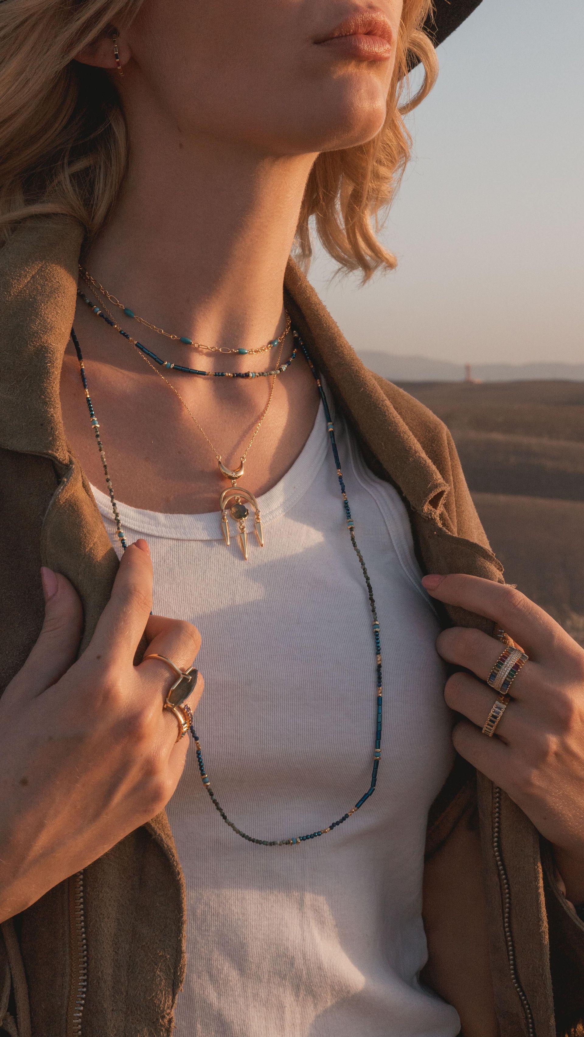 Femme portant des colliers et des bagues superposés, un haut blanc et une veste marron, debout à l'extérieur.