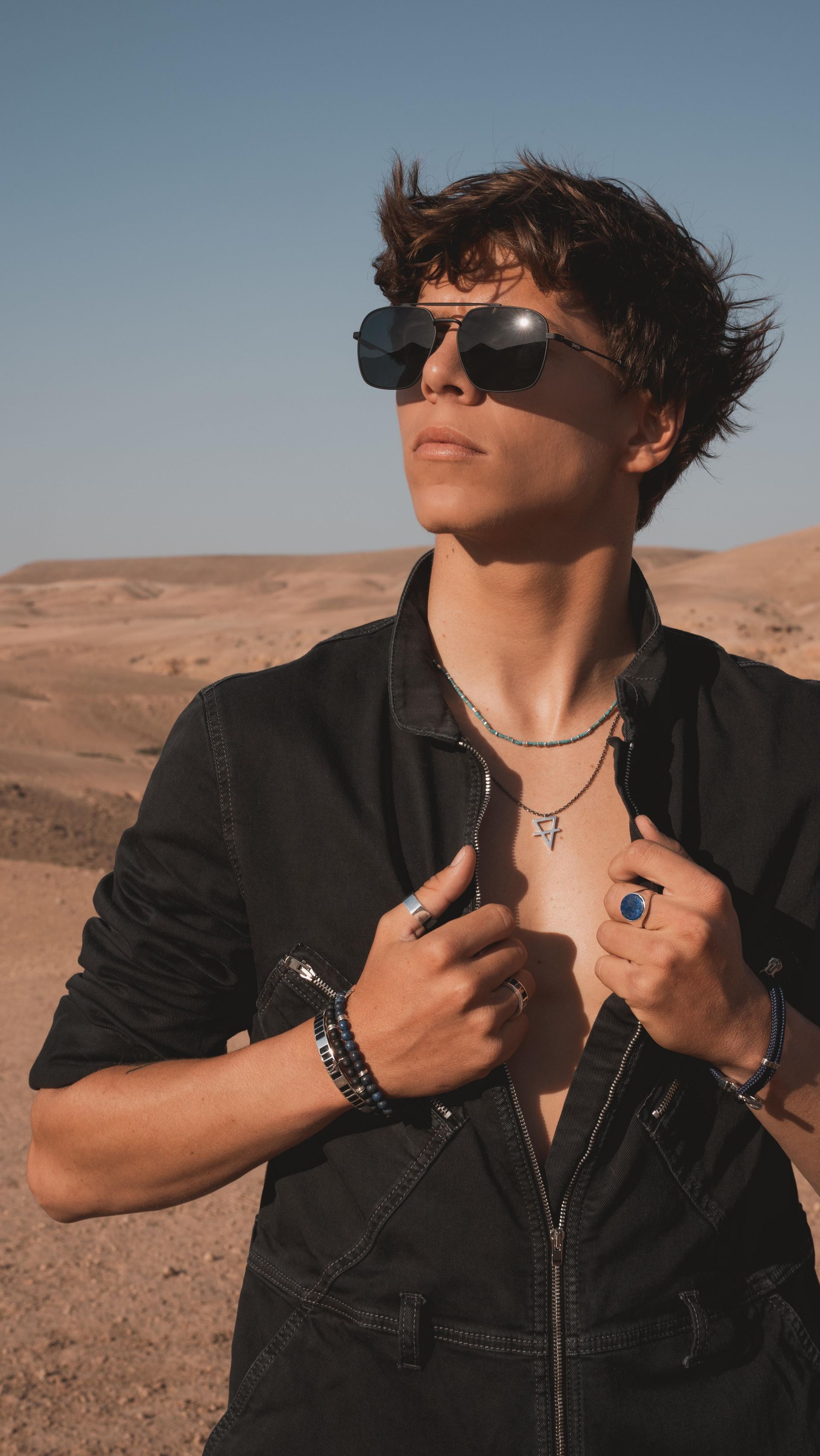 Jeune dans le désert, portant des lunettes de soleil et une tenue noire, regardant vers le haut.
