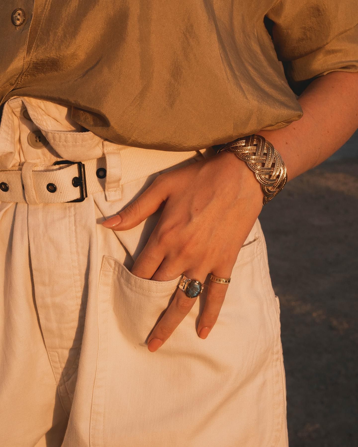 Personne en tenue beige, main dans la poche. Bracelet et bagues en or. Lumière du soleil.