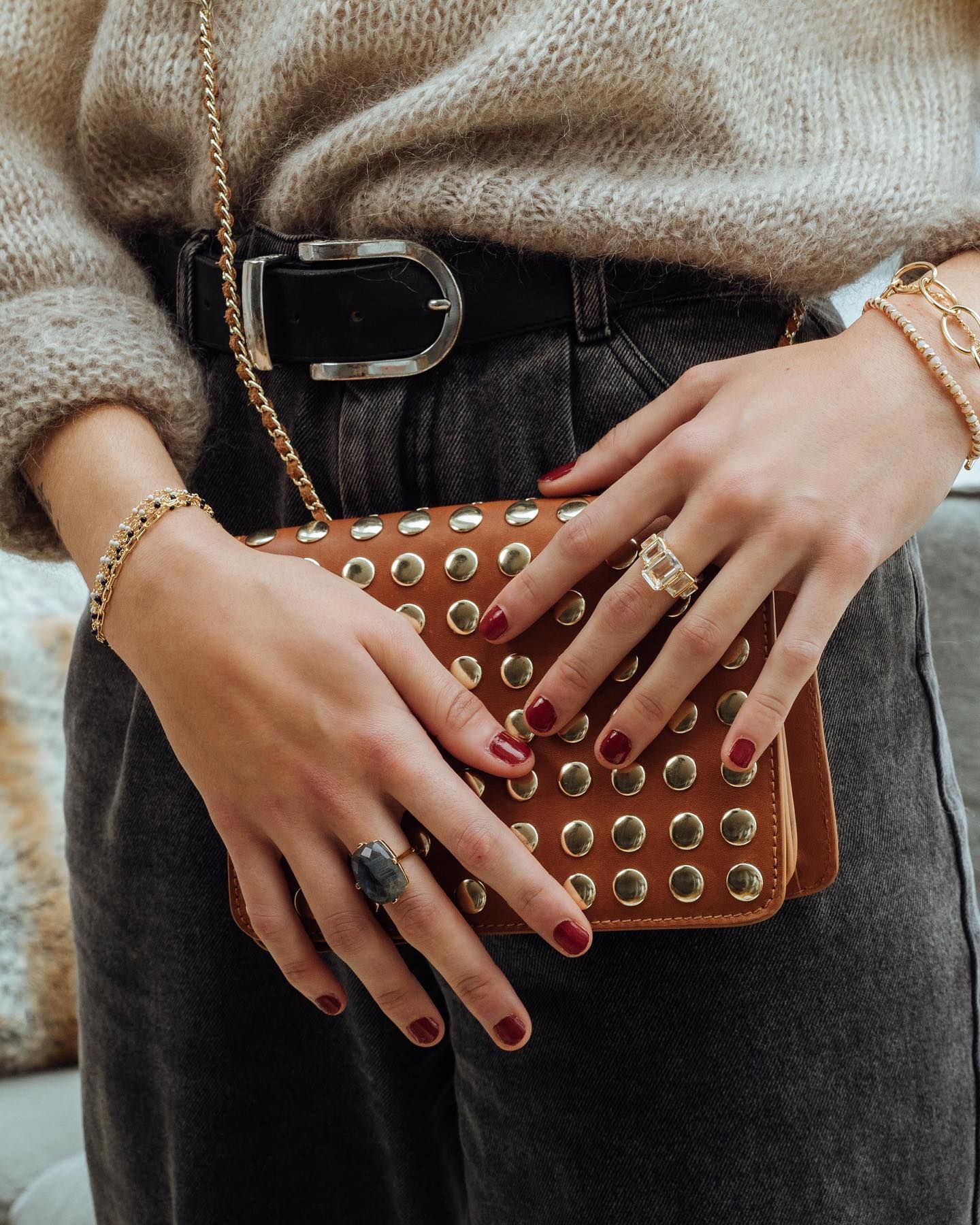 Femme en pull et jean, tenant un sac à main marron clouté, portant des bagues et des bracelets, avec du vernis à ongles rouge.
