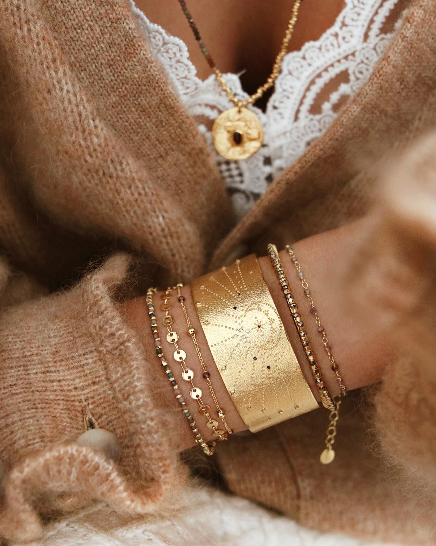 Femme portant des bijoux en or : colliers, bracelets et manchette. Pull beige et haut en dentelle blanche.