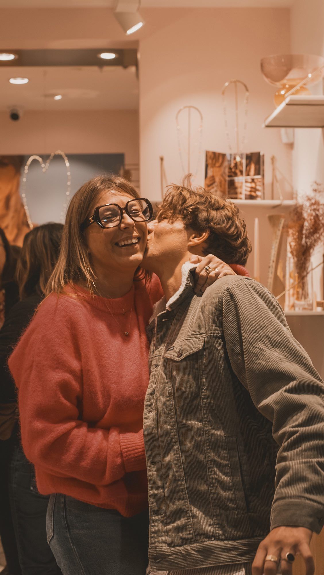 Une femme en pull rouge sourit tandis qu'un jeune homme l'embrasse sur la joue ; tons chauds, peut-être un intérieur de magasin.