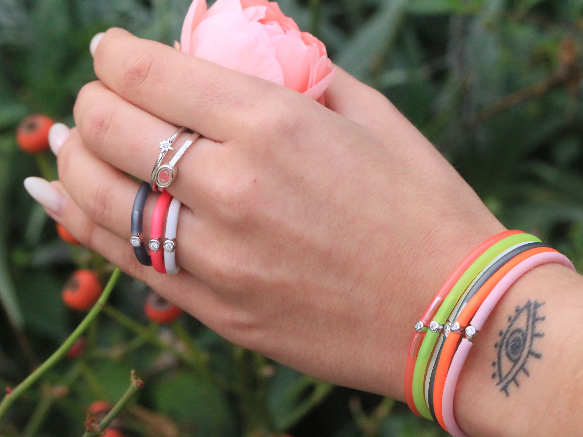 Main de femme portant bagues et bracelets tenant une fleur rose, près d'un rosier. Œil tatoué sur le poignet.