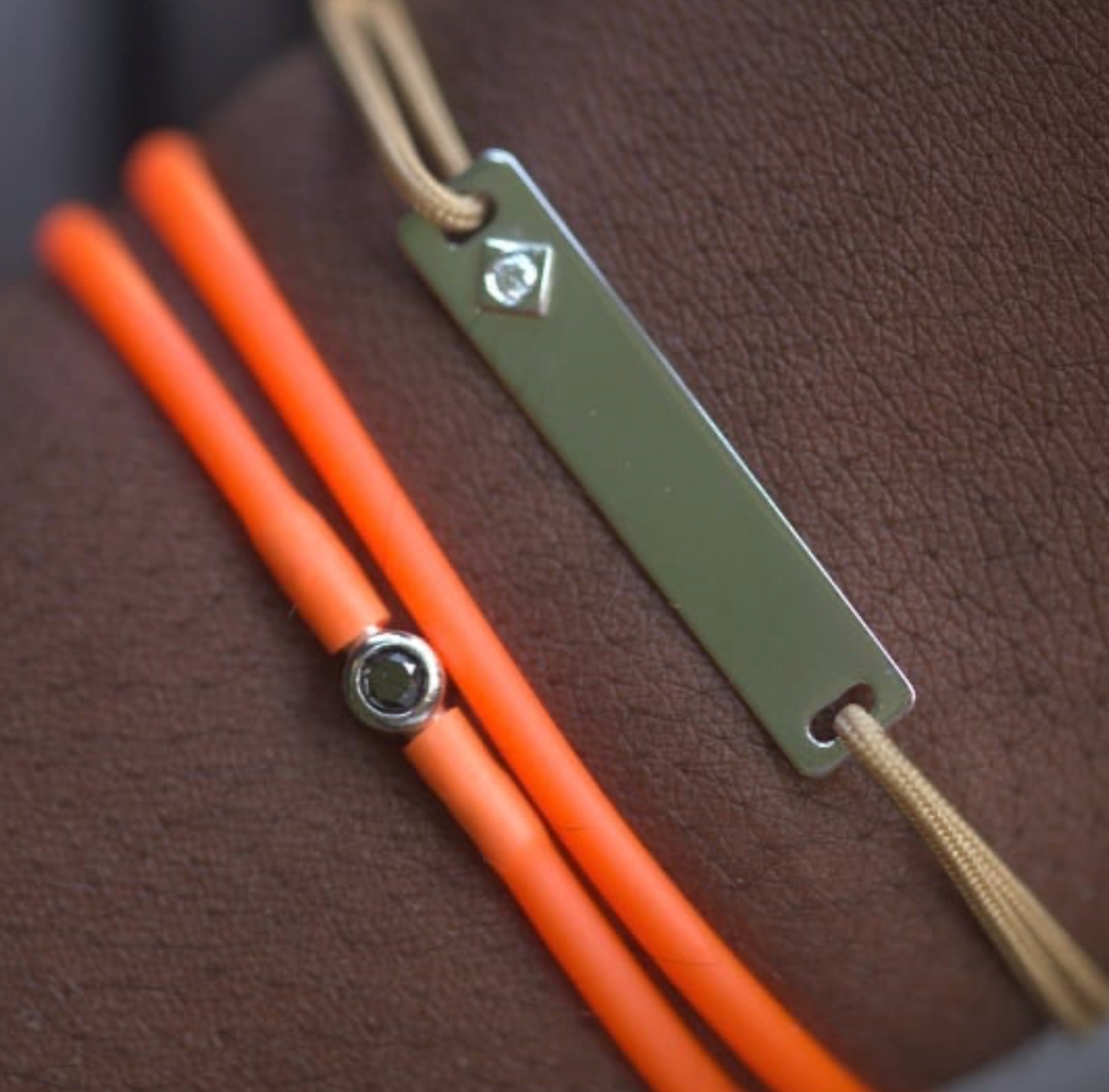 Gros plan de deux bracelets sur un bras à la peau foncée. L'un est orange, l'autre est doté d'une breloque rectangulaire argentée et d'un cordon beige.