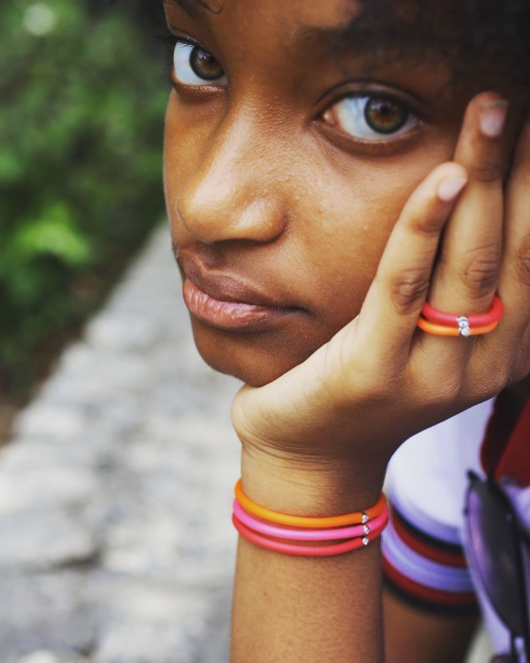 Jeune personne à la peau brune et aux yeux verts, le menton posé sur la main, portant des bagues et des bracelets colorés.