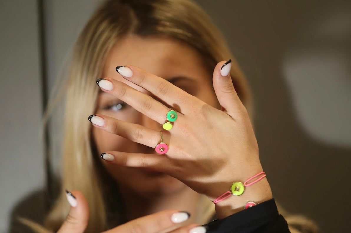 Femme blonde aux ongles à la française, exhibant une bague verte et un bracelet rose avec des breloques en forme de bouton.