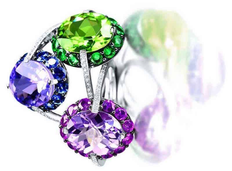 Bague avec trois grosses pierres précieuses : bleue, verte et violette, chacune entourée de pierres plus petites, fond blanc.