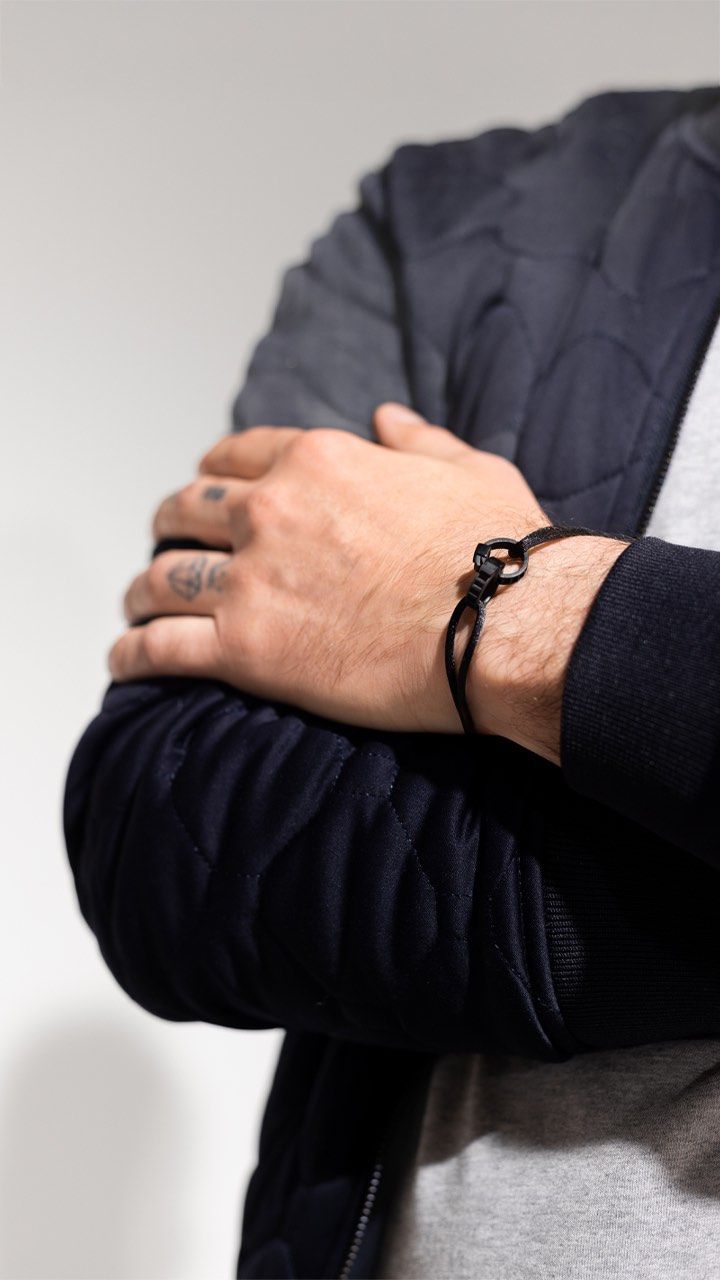 Homme portant un bracelet noir, les bras croisés, portant une veste matelassée bleue et une chemise grise.