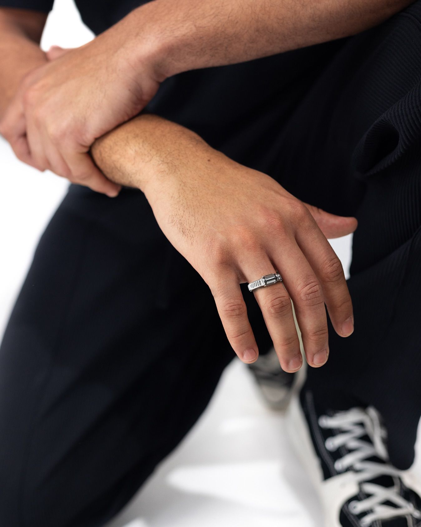 La main de l'homme, portant une bague, repose sur son autre bras. Il porte des vêtements sombres et des baskets blanches.