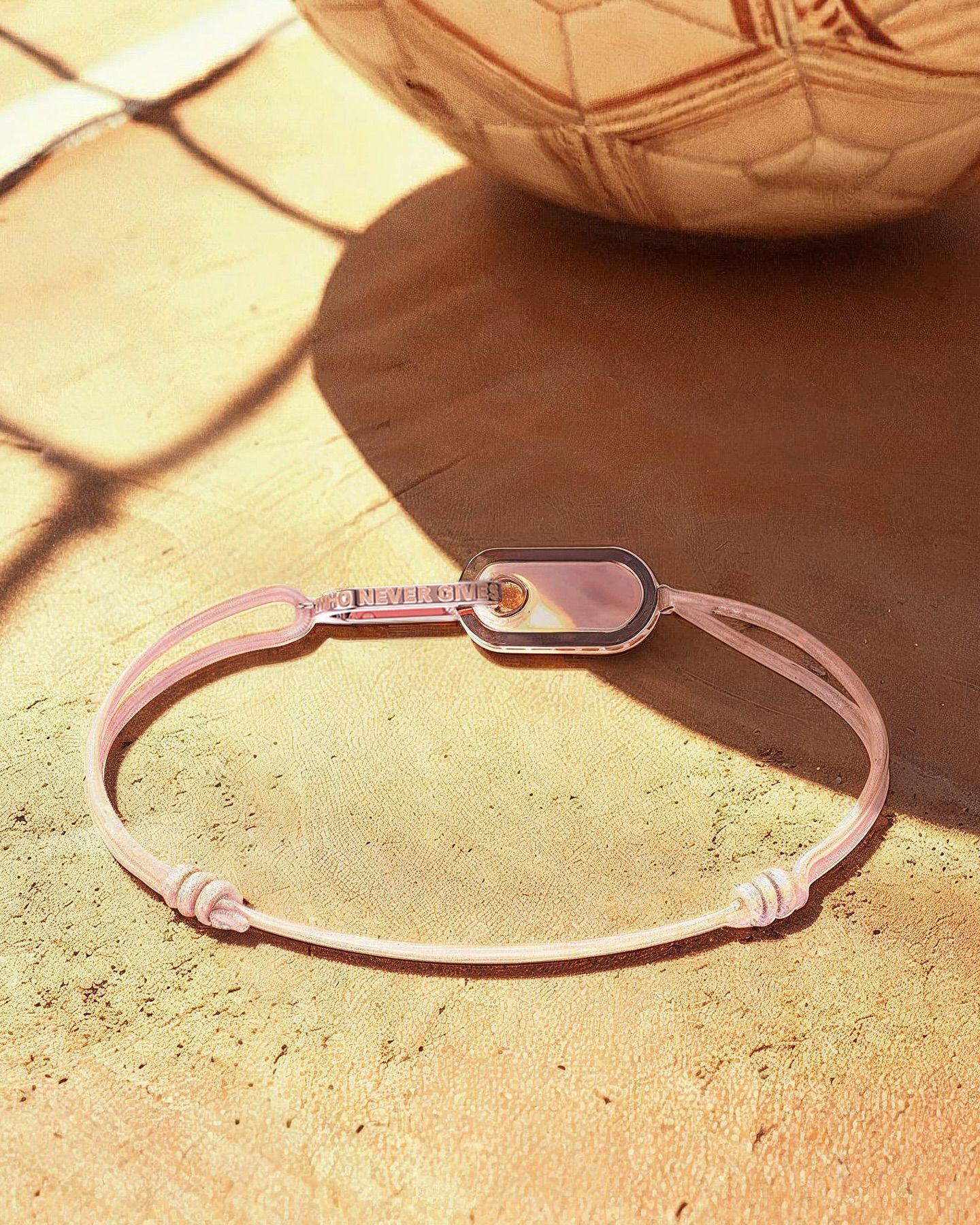 Bracelet cordon rose avec une breloque rectangulaire, sur une surface sablonneuse.