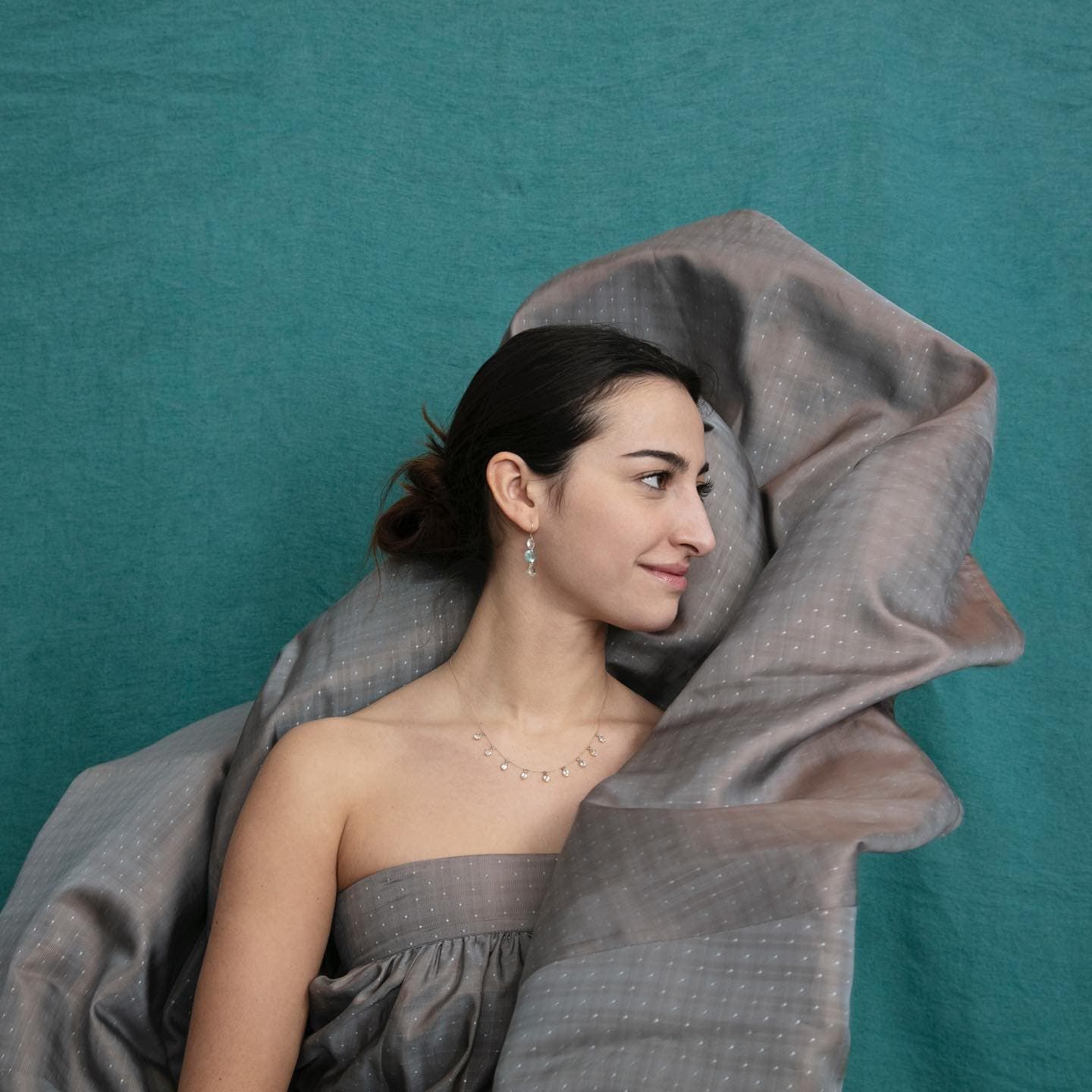 Femme en robe grise sans bretelles avec du tissu flottant autour d'elle ; fond turquoise.