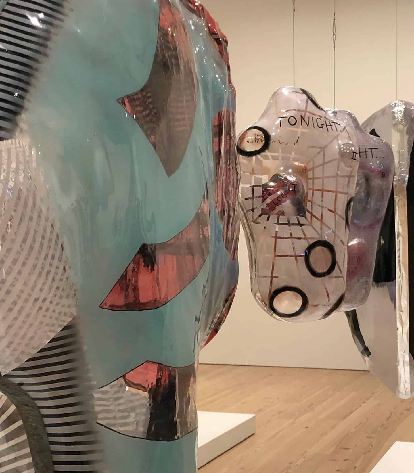 Sculptures abstraites dans une galerie. Forme turquoise aux accents argentés et roses. Tête transparente avec texte.