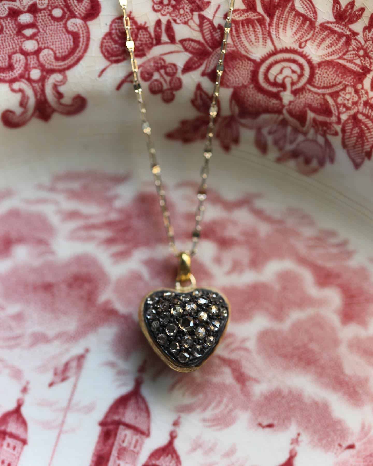 Collier en or avec un pendentif en forme de cœur recouvert de pierres sombres, reposant sur une surface à motifs rouges et blancs.