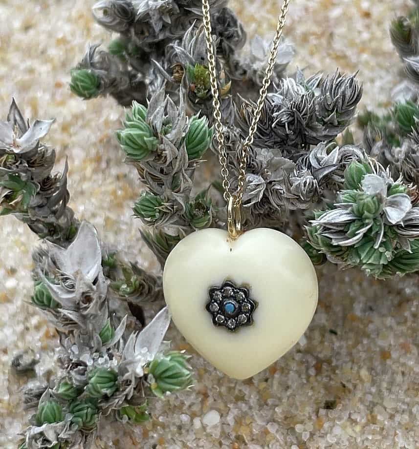 Pendentif en forme de coeur crème avec centre bleu, sur chaîne dorée, reposant sur des plantes vertes et grises, fond sable.