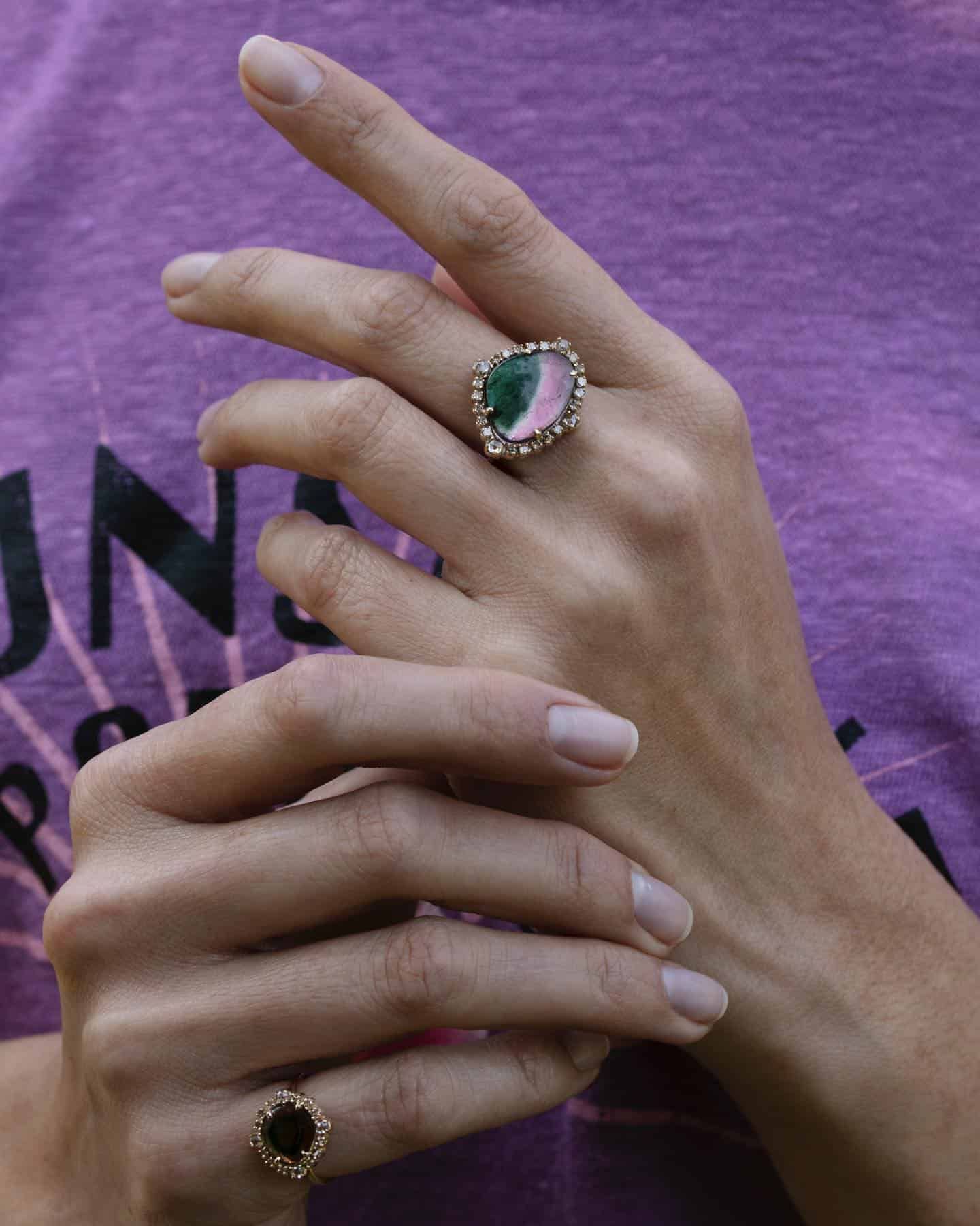 Main portant une grande bague ornée d'une pierre rose et verte. L'autre main est en dessous.