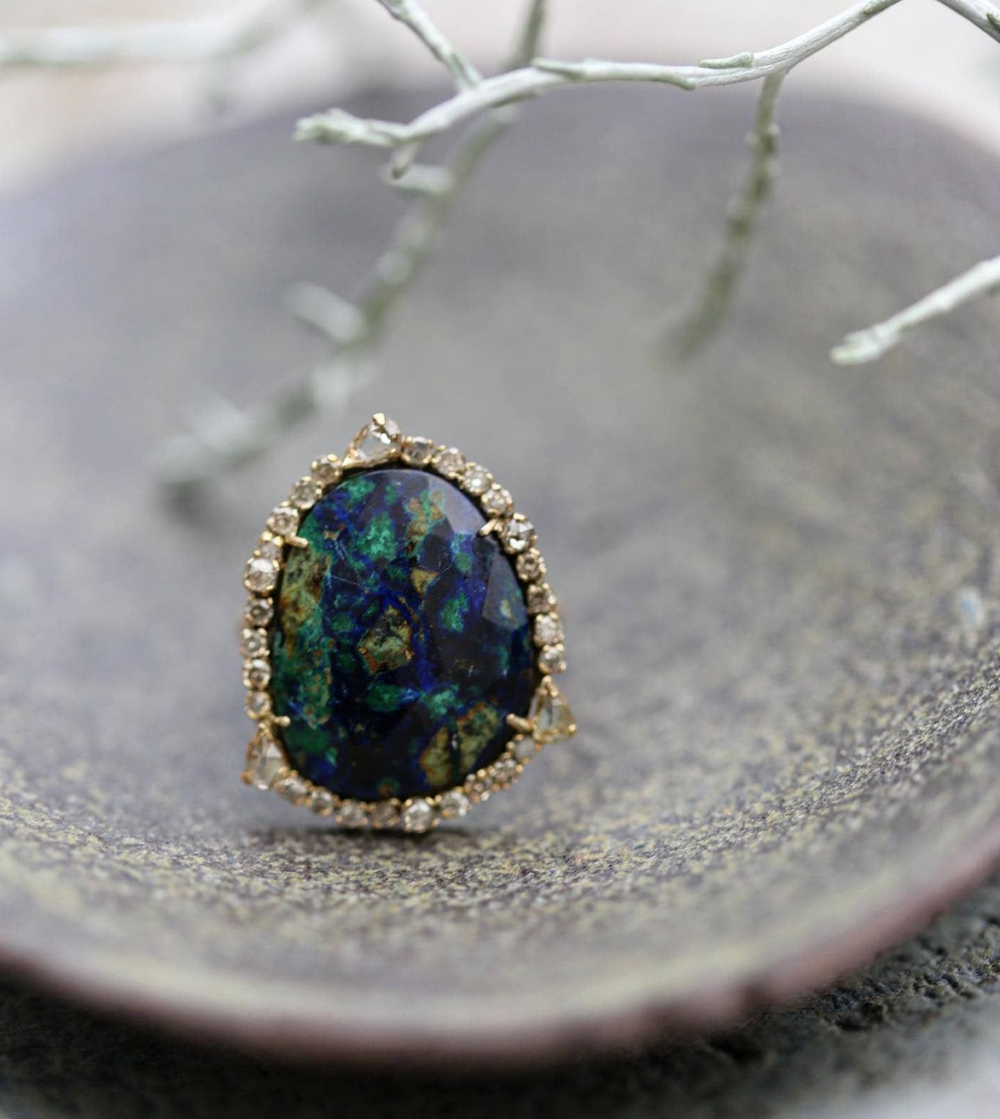 Bague en or avec une grosse pierre précieuse bleu-vert foncé entourée de petits diamants, reposant sur une surface grise.