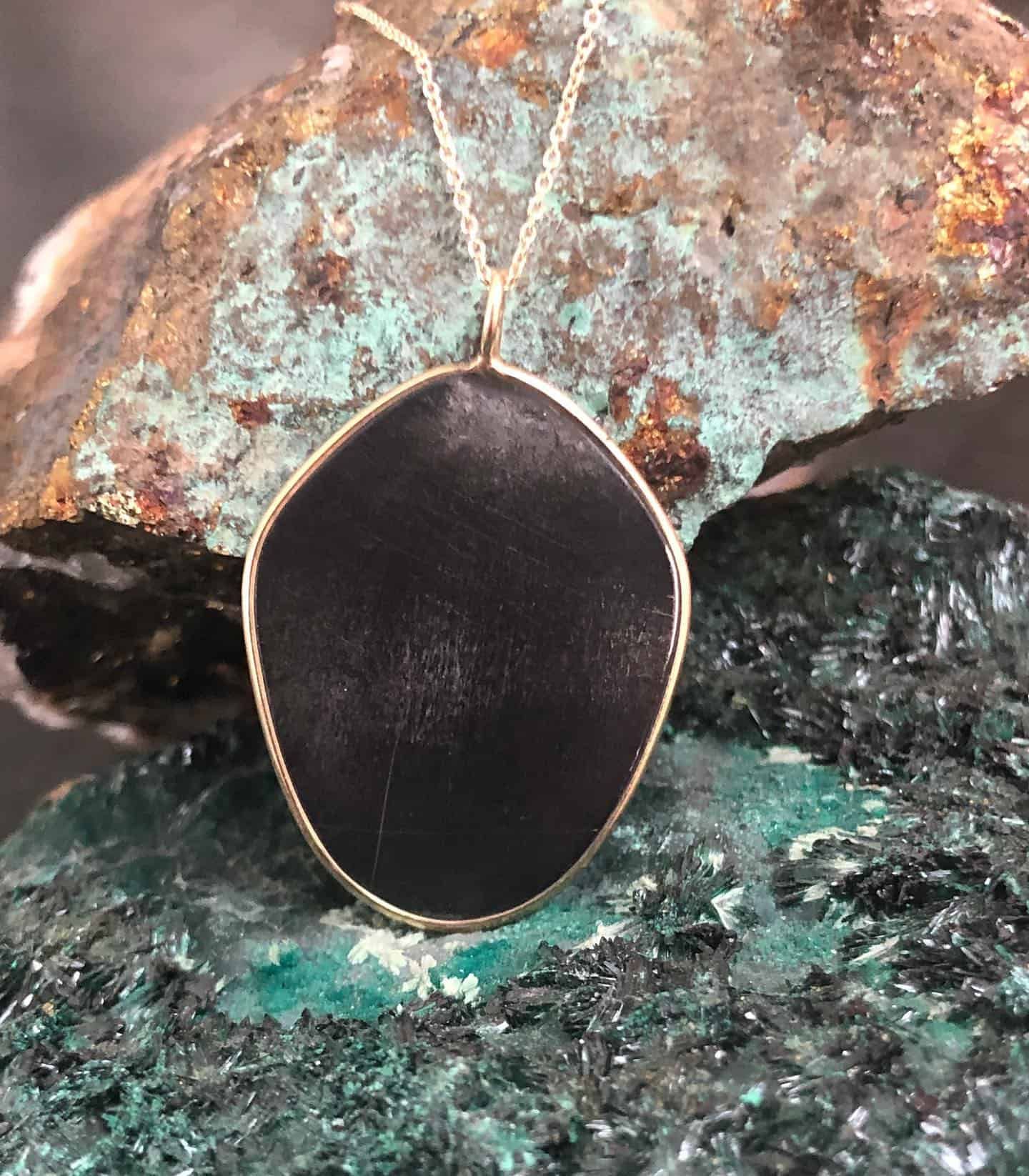 Collier pendentif noir sur une chaîne en or, sur fond de roches texturées vertes et brunes.