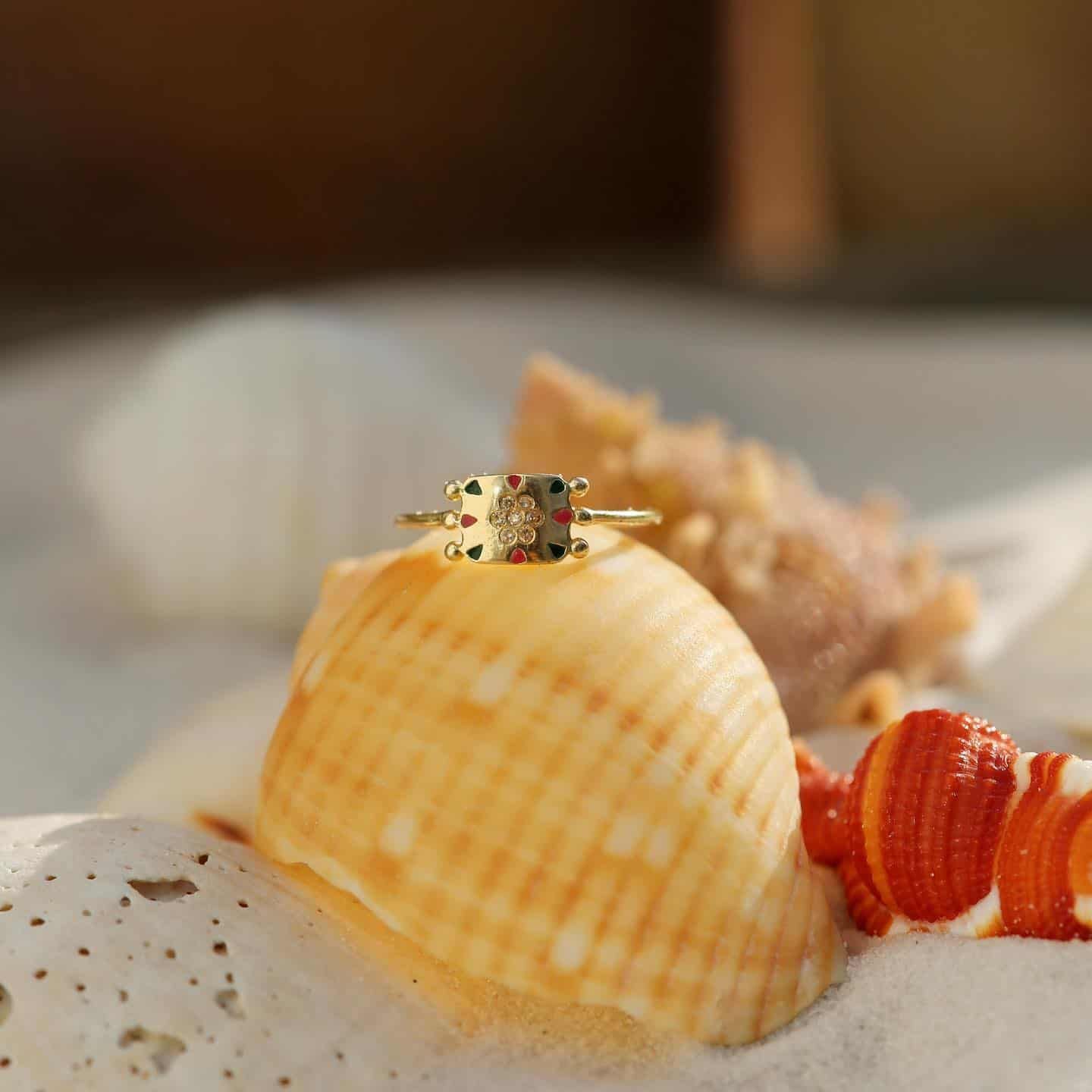 Bague en or avec motif de carte à jouer reposant sur un coquillage avec d'autres coquillages en arrière-plan.